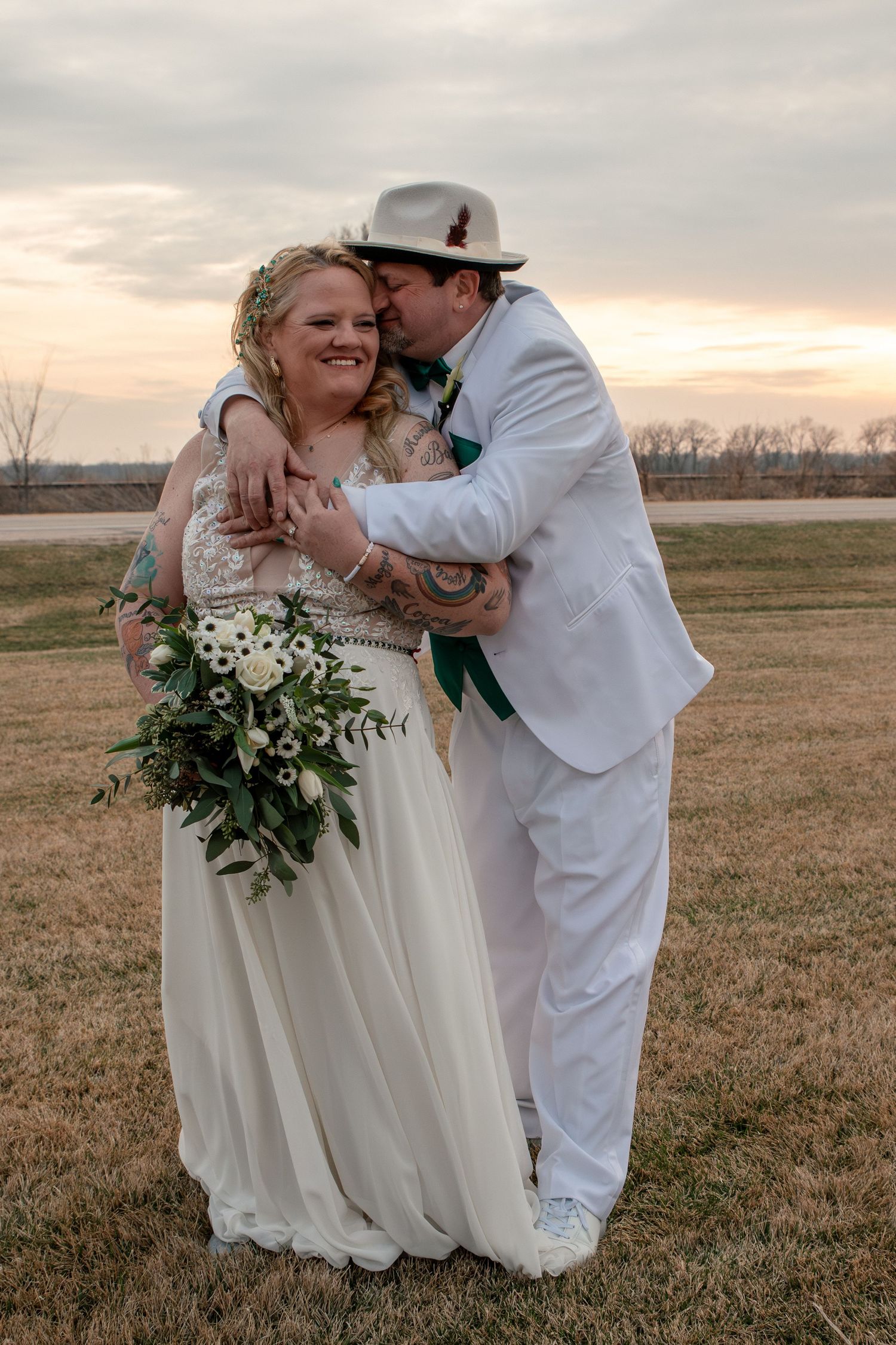 A groom in a white suit and hat kisses his smiling bride holding a lush green bouquet at sunset.