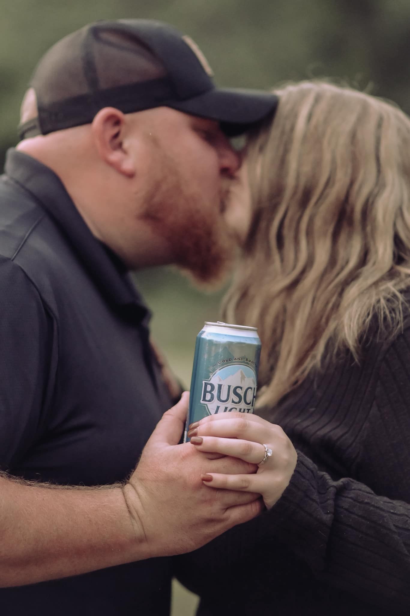 Engaged couple kissing while holding a Busch Light beer can, showcasing a diamond engagement ring outdoors.