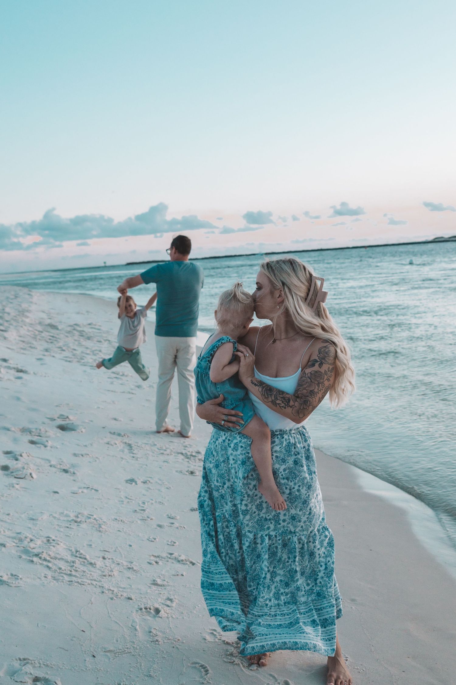 Family enjoying a beach sunset, woman with tattoos in teal maxi dress kissing toddler, man playing with child by ocean.