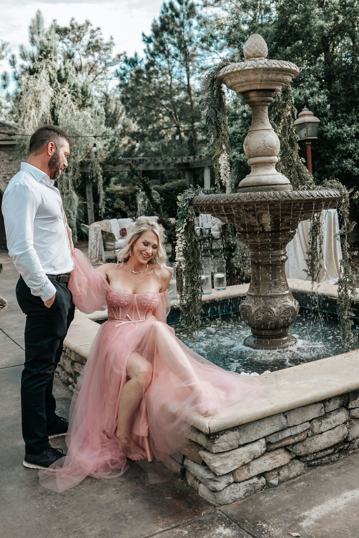 Woman in elegant pink tulle gown sits on stone fountain edge while man in white shirt smiles at her.