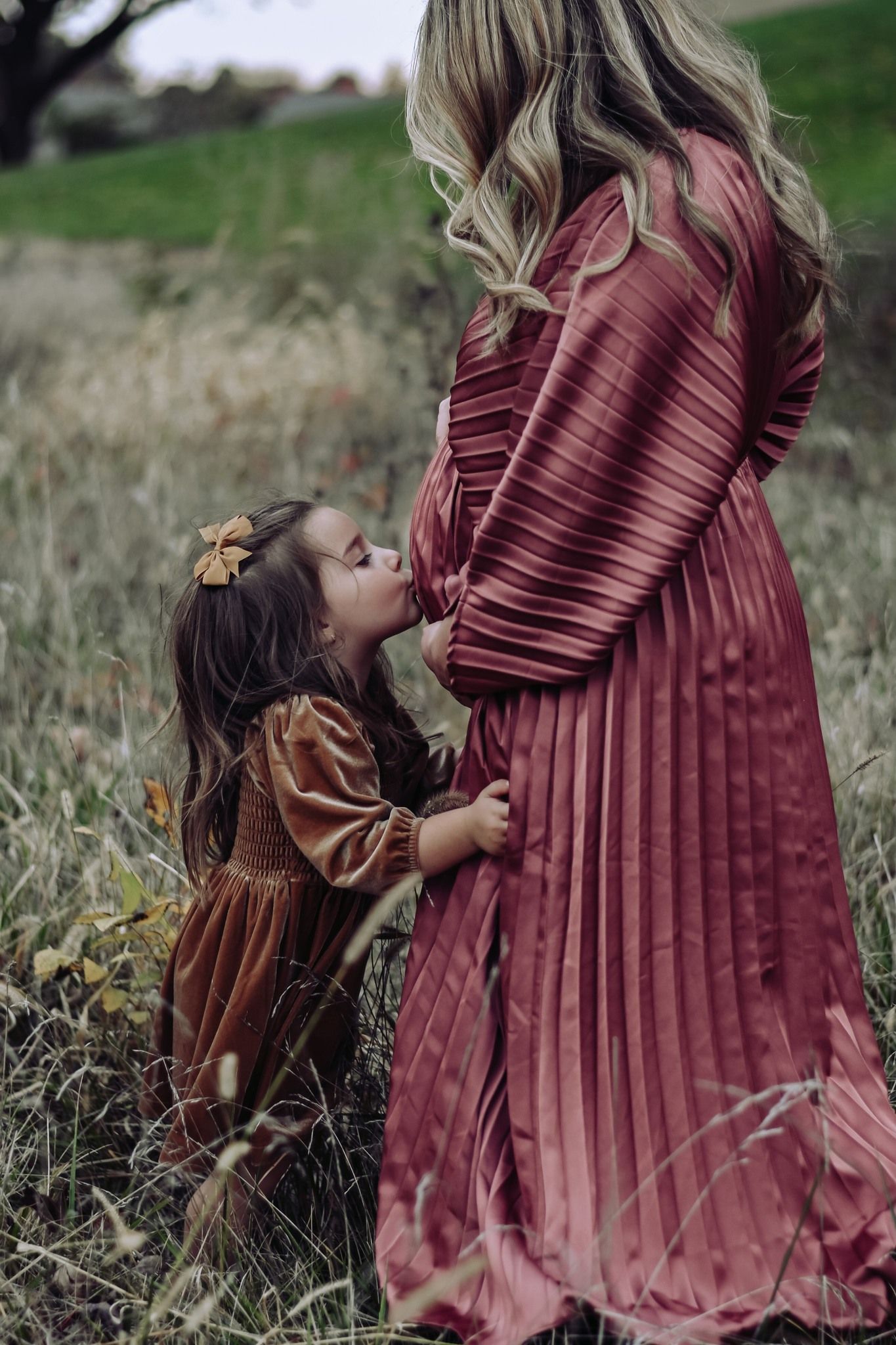A little girl in a brown dress kisses her pregnant mother's belly in a field during autumn.