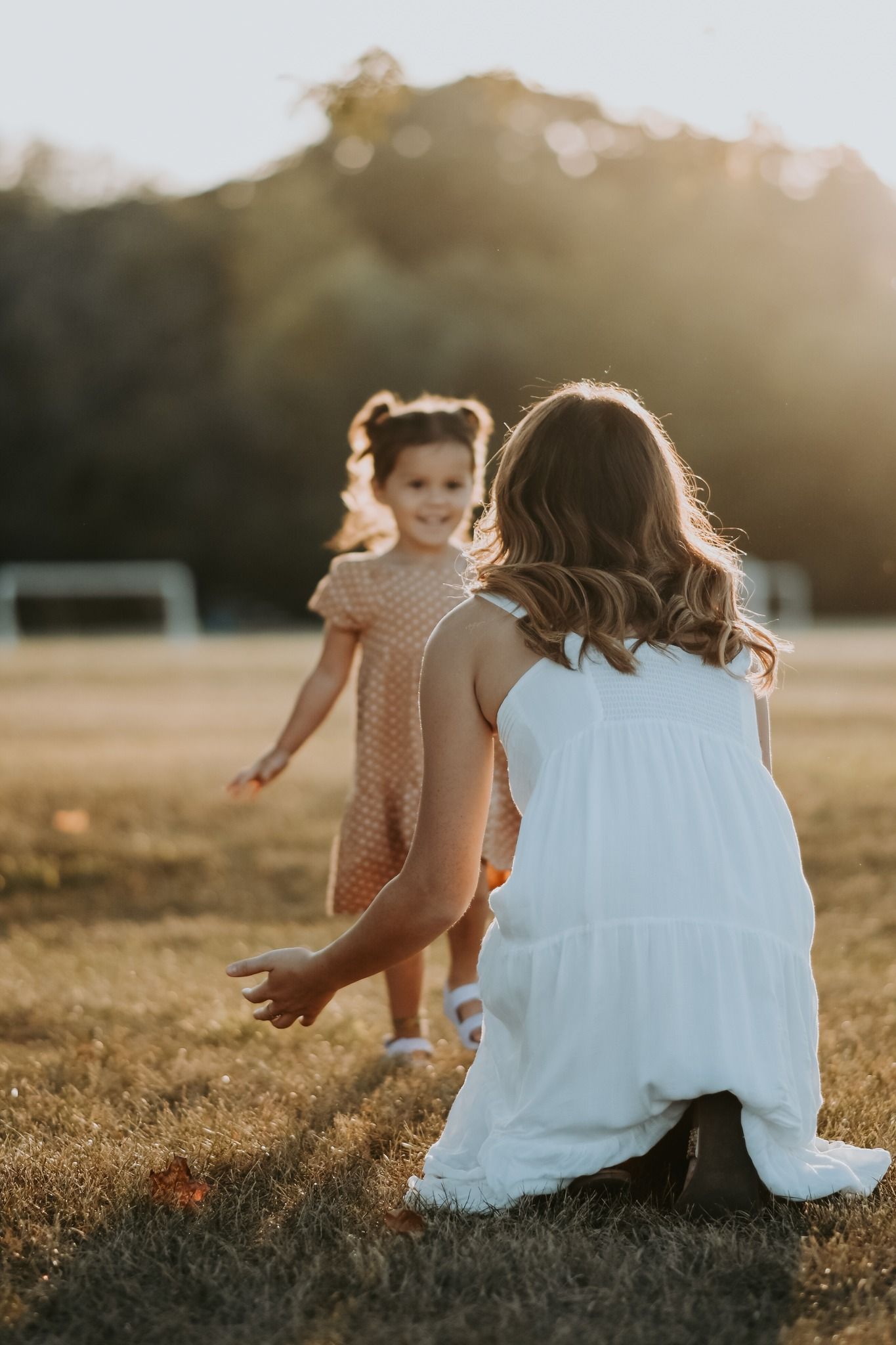 A mother kneels in golden sunlight reaching toward her smiling toddler daughter in a pink dress.