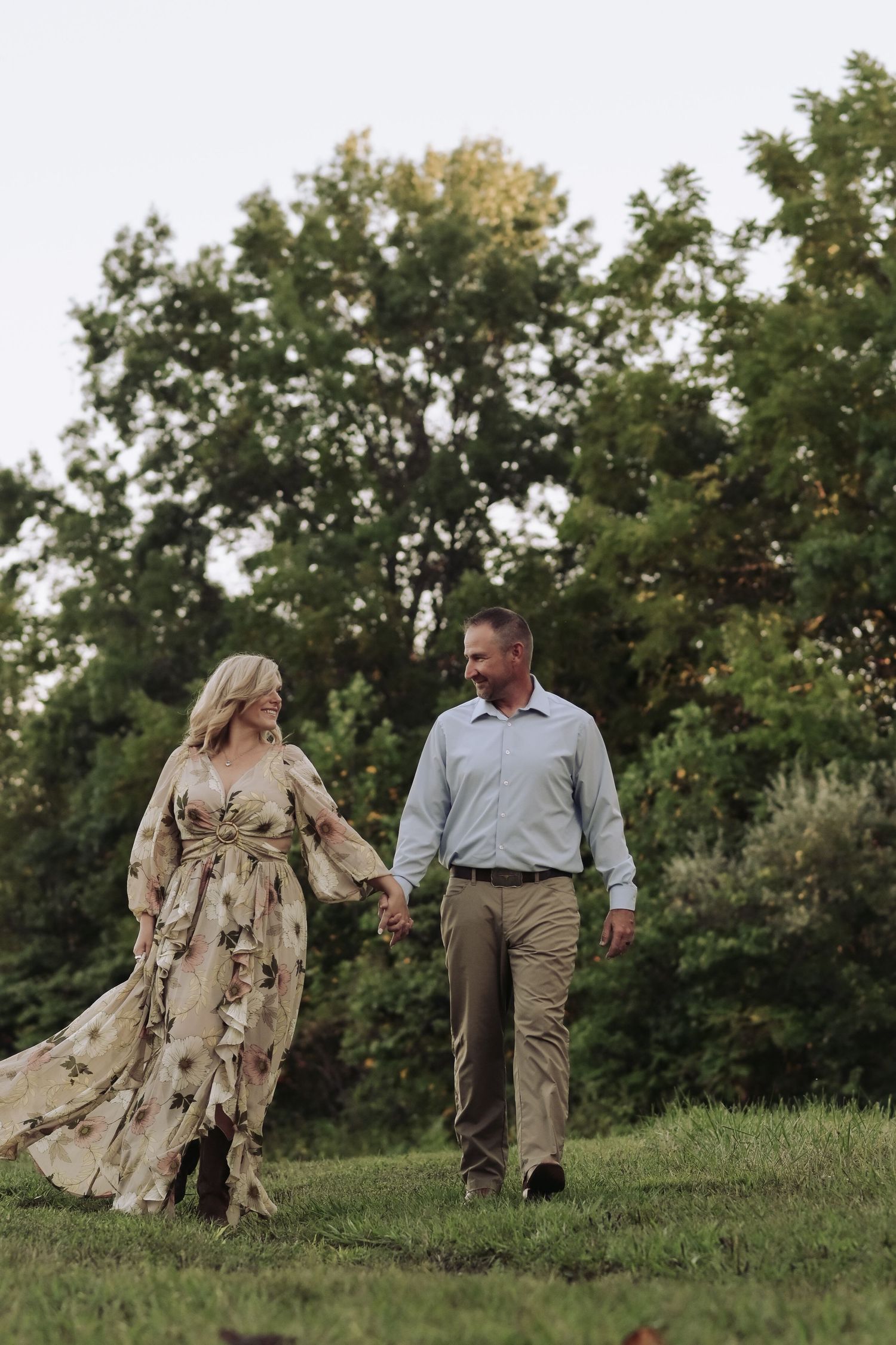 Happy couple walking and laughing together on green lawn, woman in flowing floral dress, trees behind.