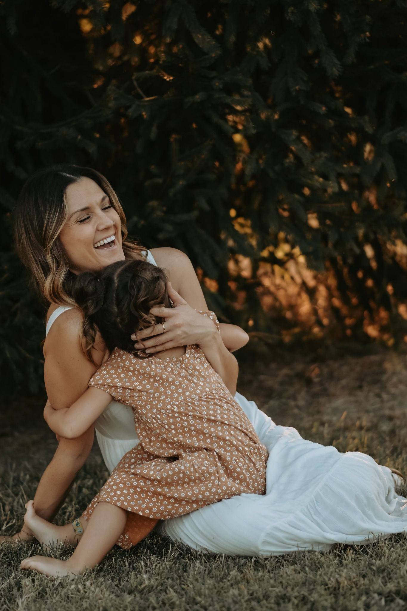 Joyful mother laughing while embracing her young daughter on grass in front of lush green trees.