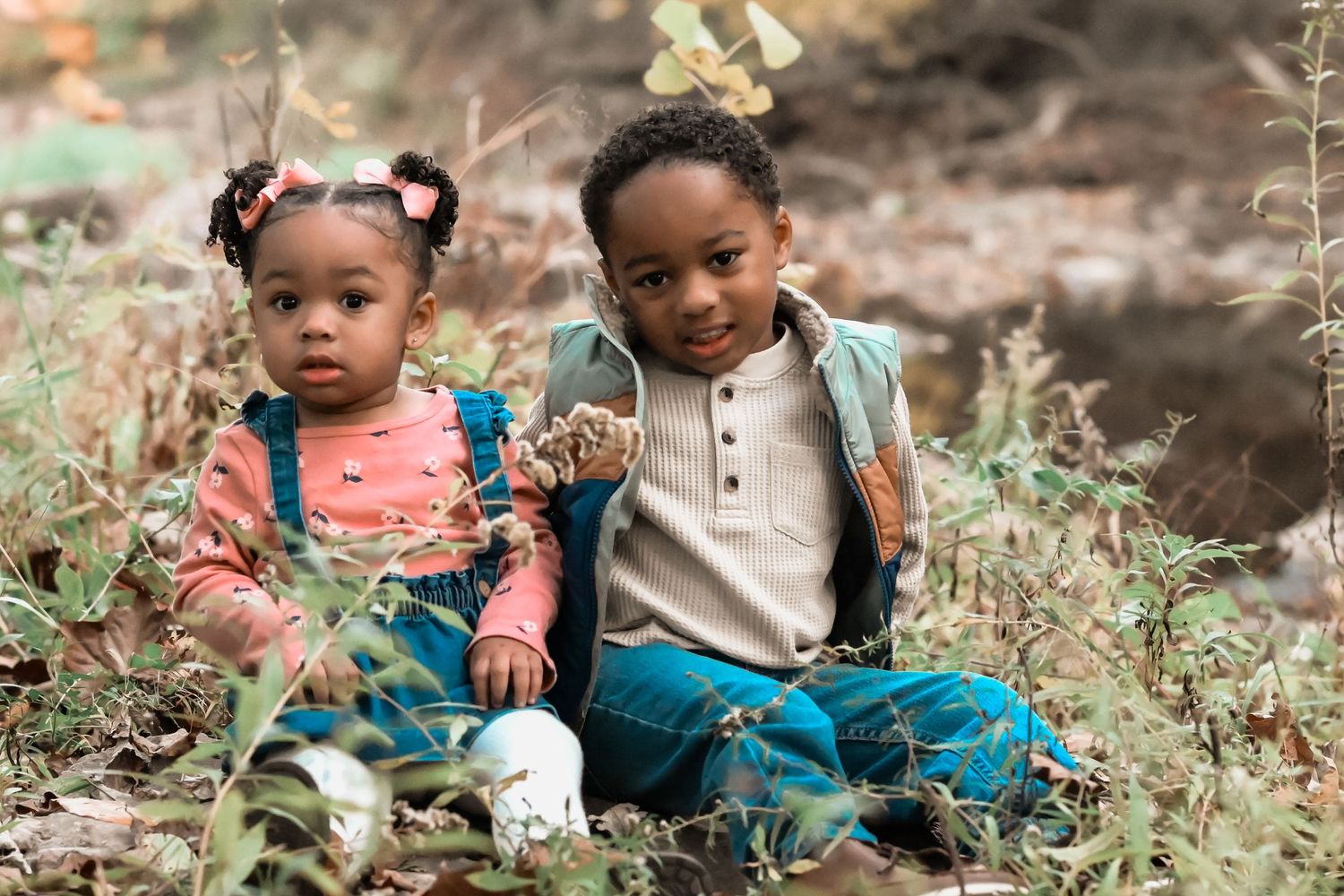 Two young Black siblings sitting together in autumn foliage outdoors.