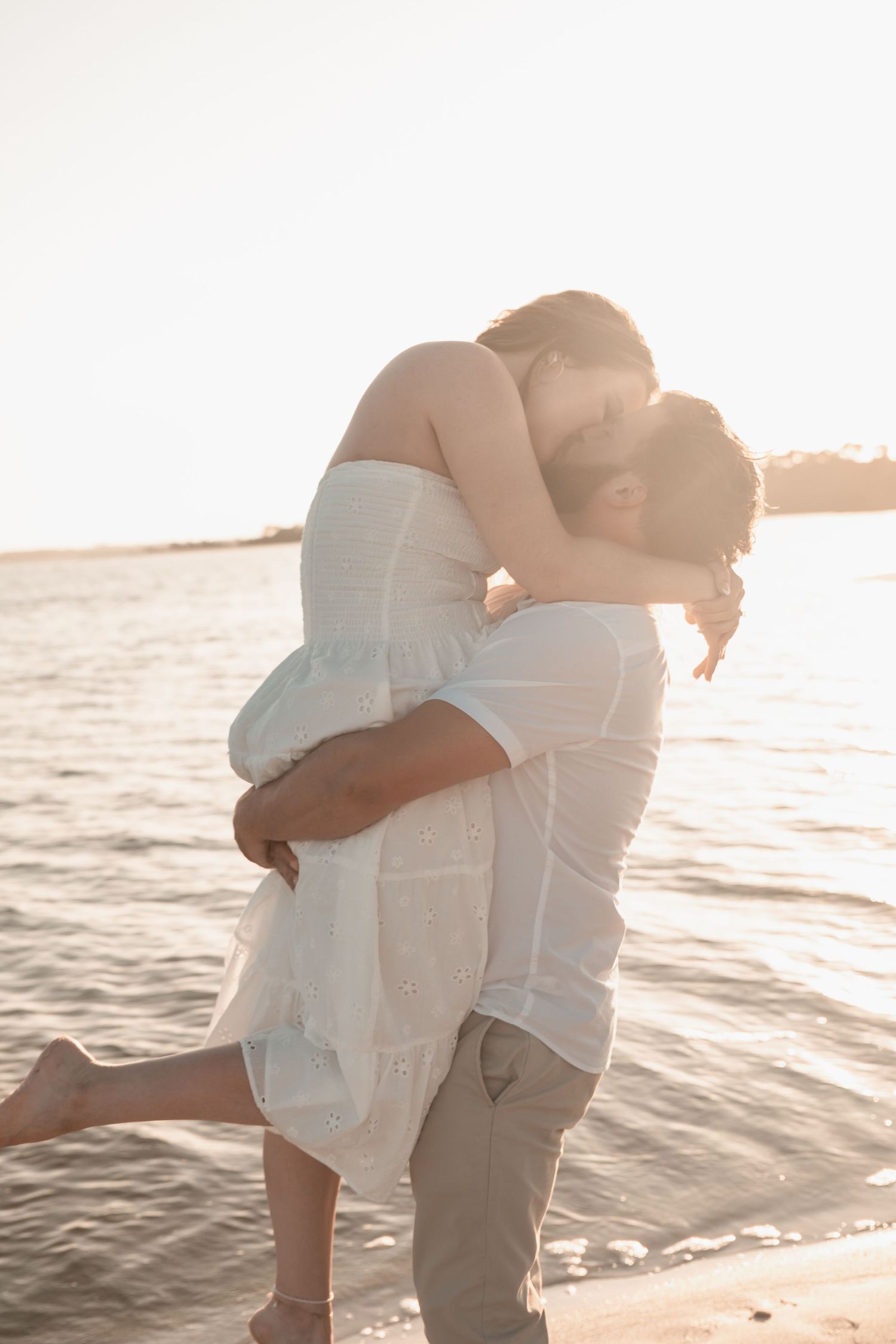 Man lifting woman in white dress for a kiss on the beach at golden hour by the water.