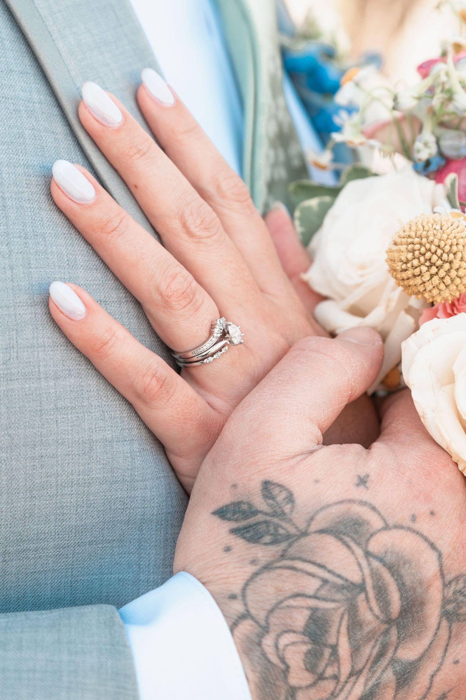 Close-up of bride's wedding ring set on hand resting on groom's gray suit, beside a floral bouquet, groom has rose tattoo.