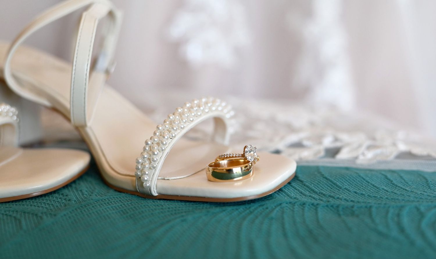 Elegant bridal heels adorned with pearls displayed alongside gold wedding bands on a teal fabric surface.