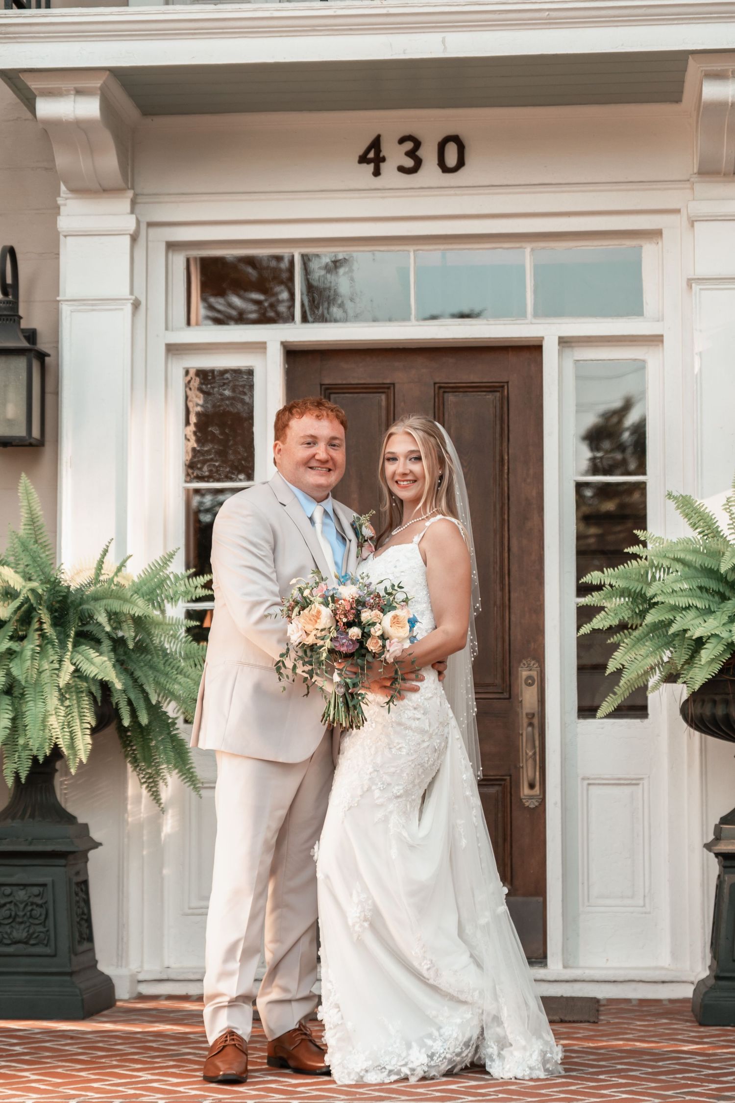 Bride and groom smiling in front of address 430, she holds a peach bouquet, he wears a tan suit with blue tie.