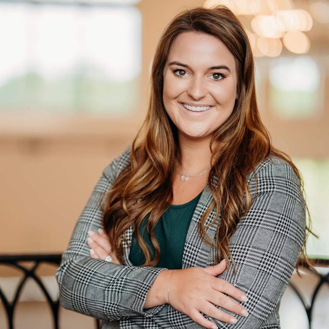 Smiling young woman in a plaid blazer poses with arms crossed in an elegant event venue setting.