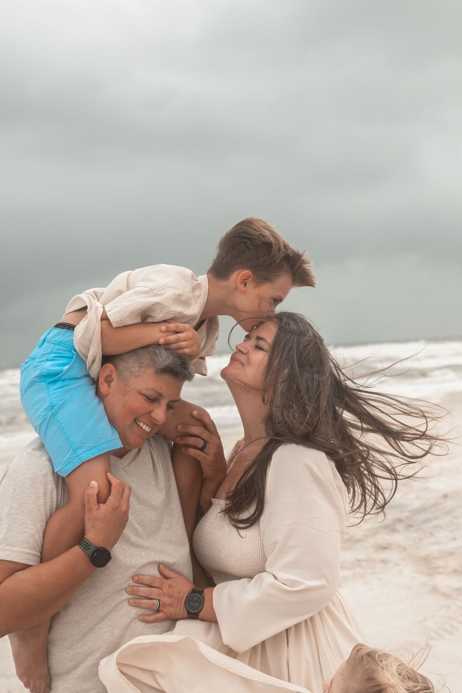 Family of three enjoying a cloudy beach day, with son on father's shoulders as mother smiles warmly nearby.
