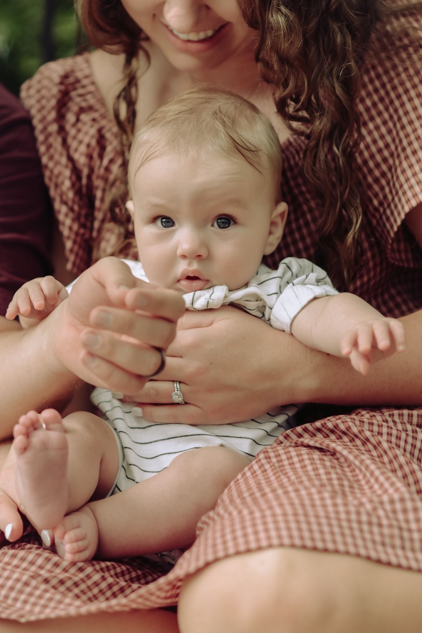 A mother in a plaid dress lovingly holds her alert baby boy on her lap outdoors.