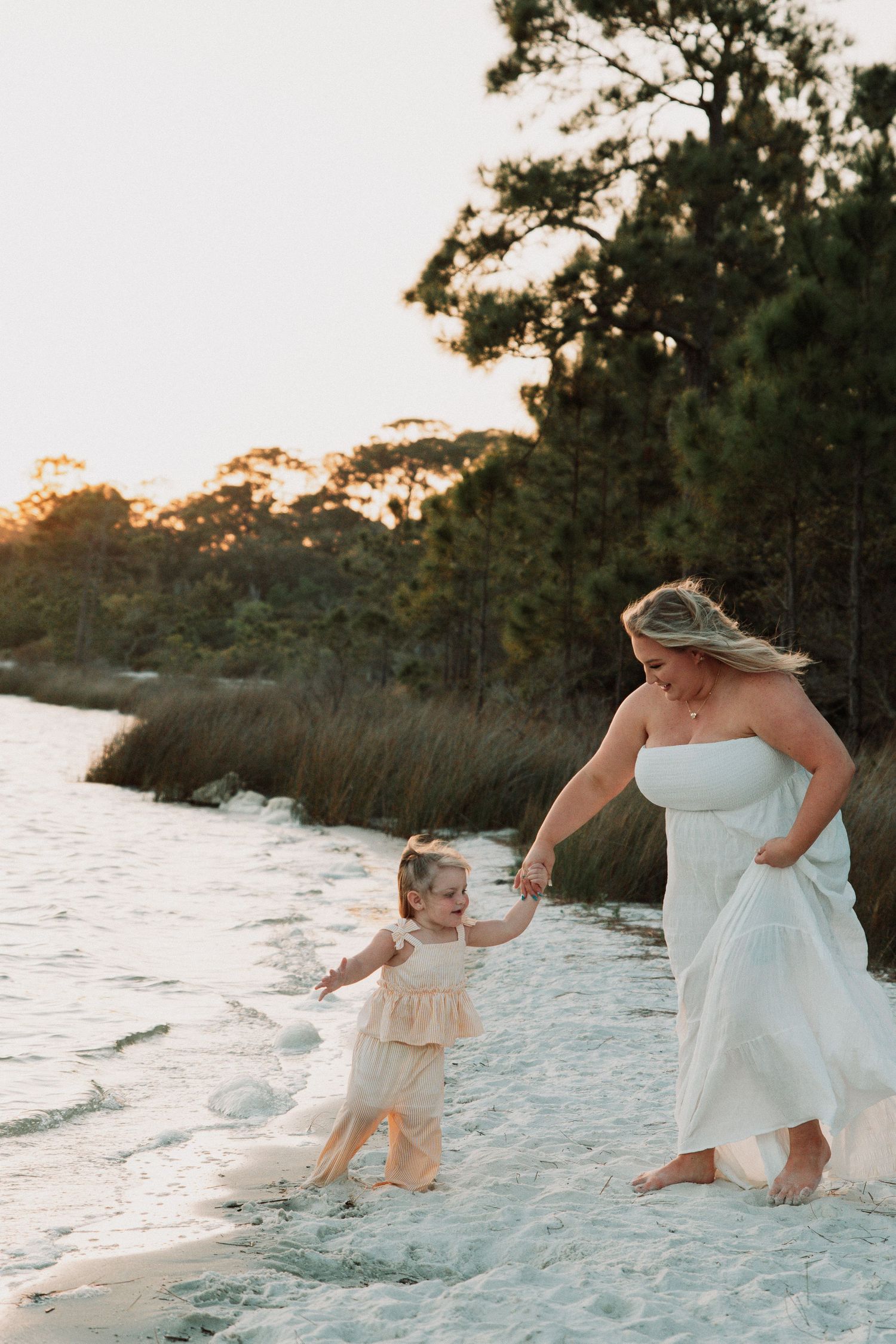 Mother in a white strapless dress holds hands with her toddler daughter while walking along a sunlit beach.