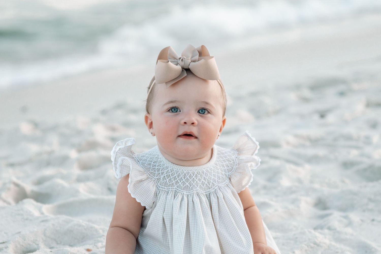 Adorable baby girl in white smocked dress and bow headband sitting on white sand beach near ocean waves.