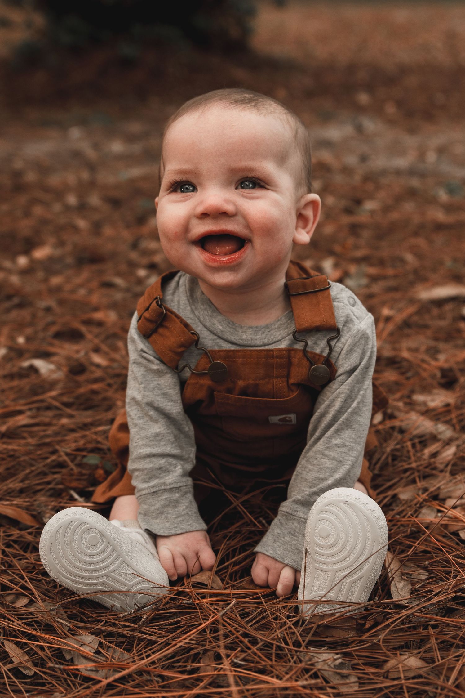 Happy baby in brown overalls sits smiling on pine needle covered ground outdoors.