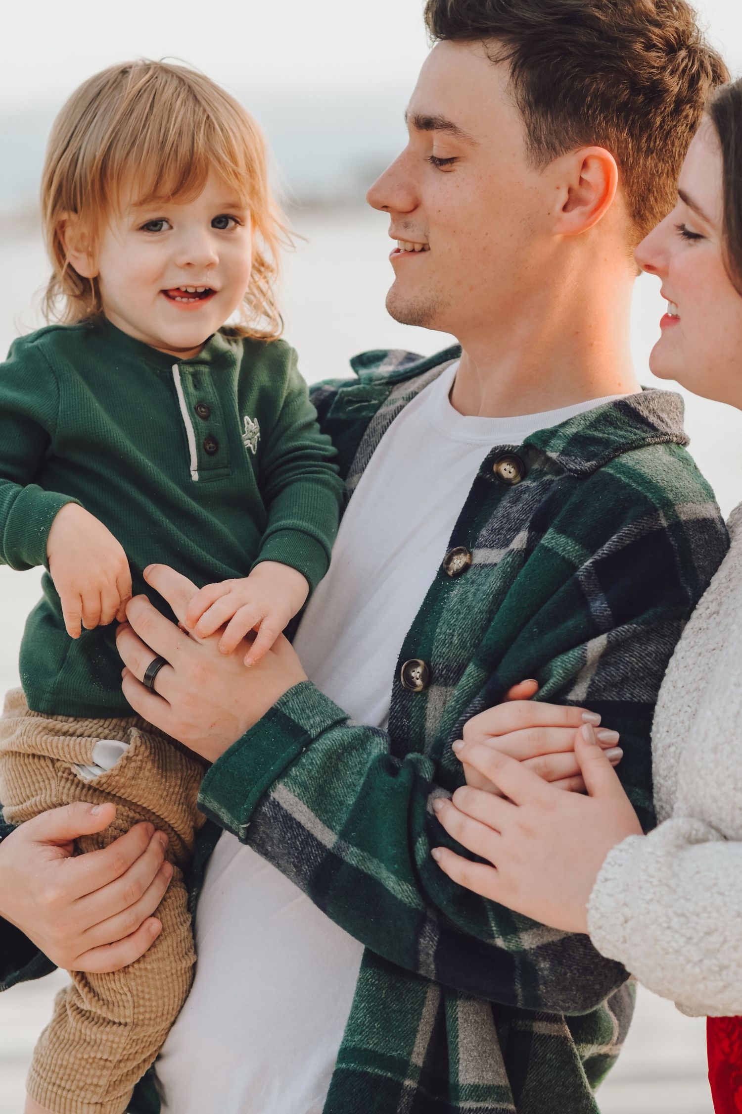 A young family smiles outdoors, father holding toddler in green outfit while mother leans in close.