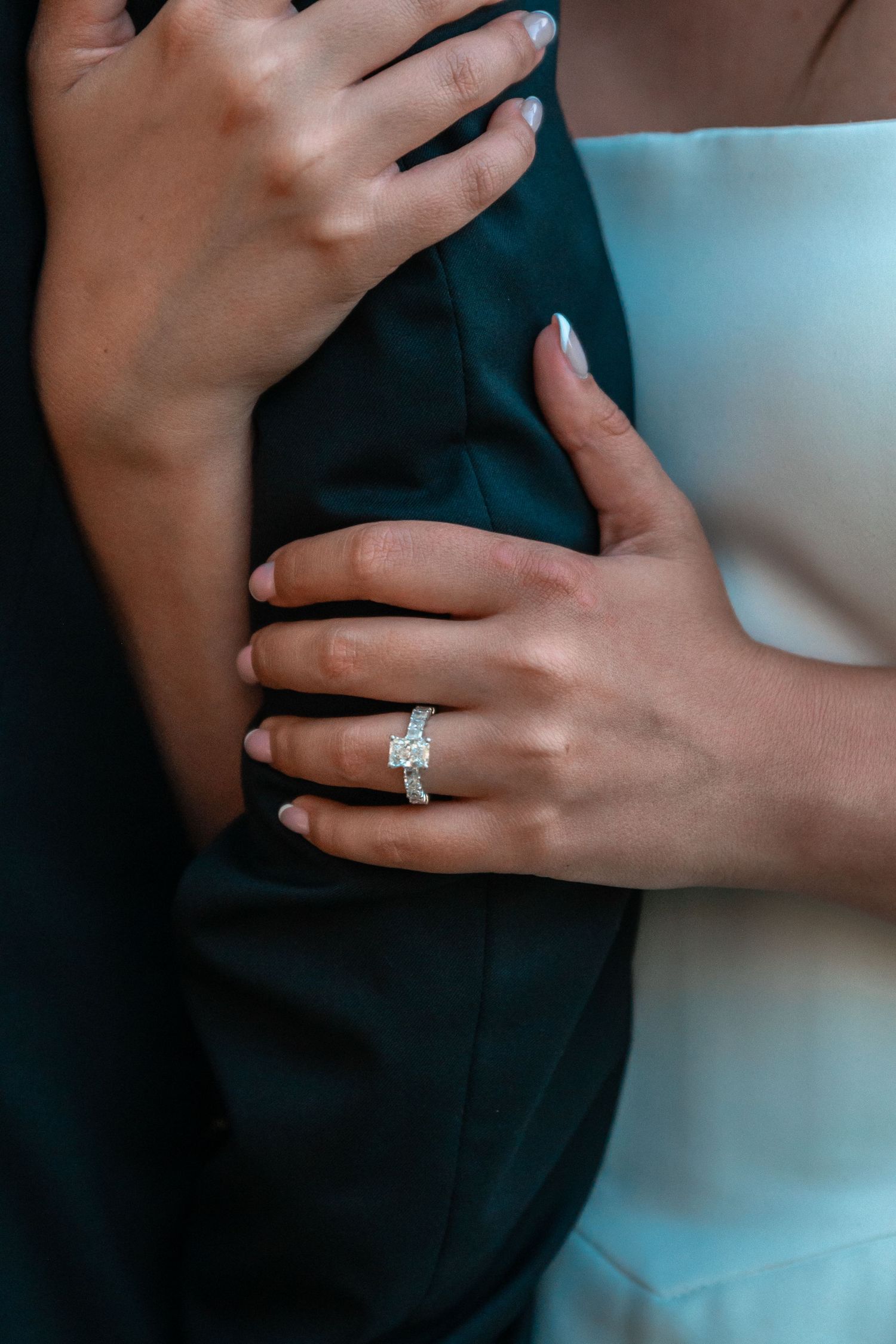 Close-up of a woman's hand adorned with a diamond engagement ring while embracing a man in a dark suit.
