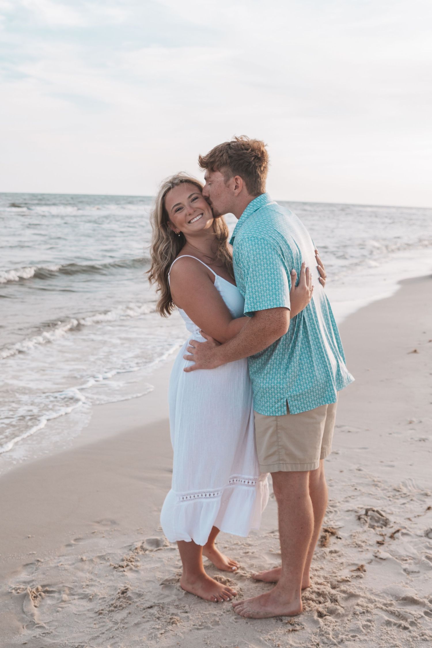 Couple embracing on a sandy beach at sunset, woman in white dress smiling as man kisses her cheek.