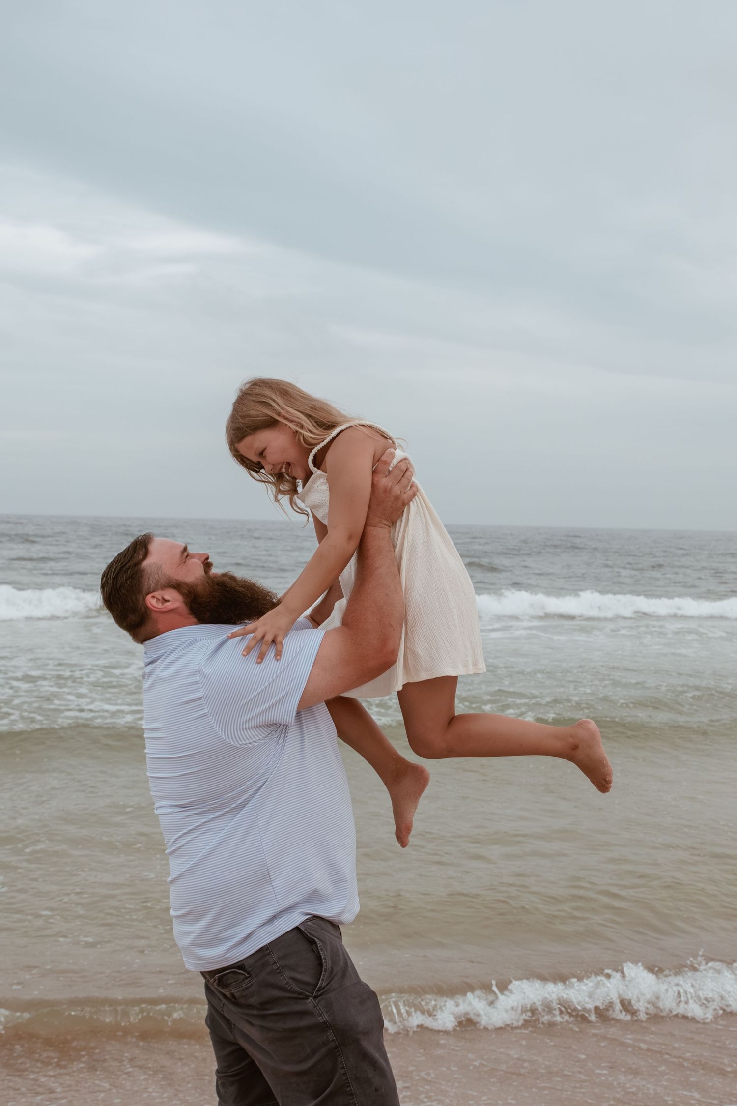 A bearded man lifts a smiling woman in a white dress on a cloudy beach with ocean waves in the background.