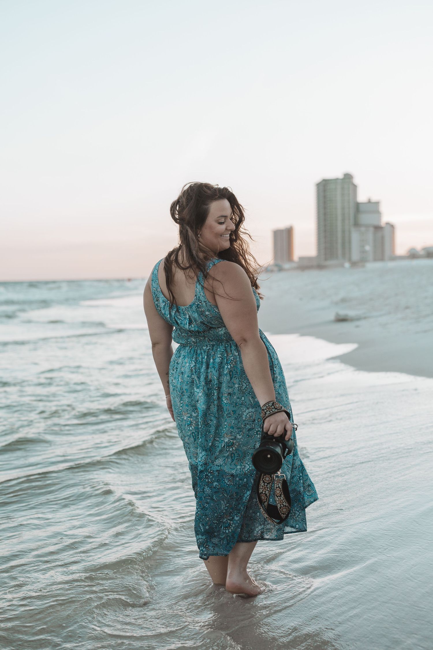 Woman in teal floral maxi dress walking barefoot on beach at sunset, holding sandals with city skyline in background.
