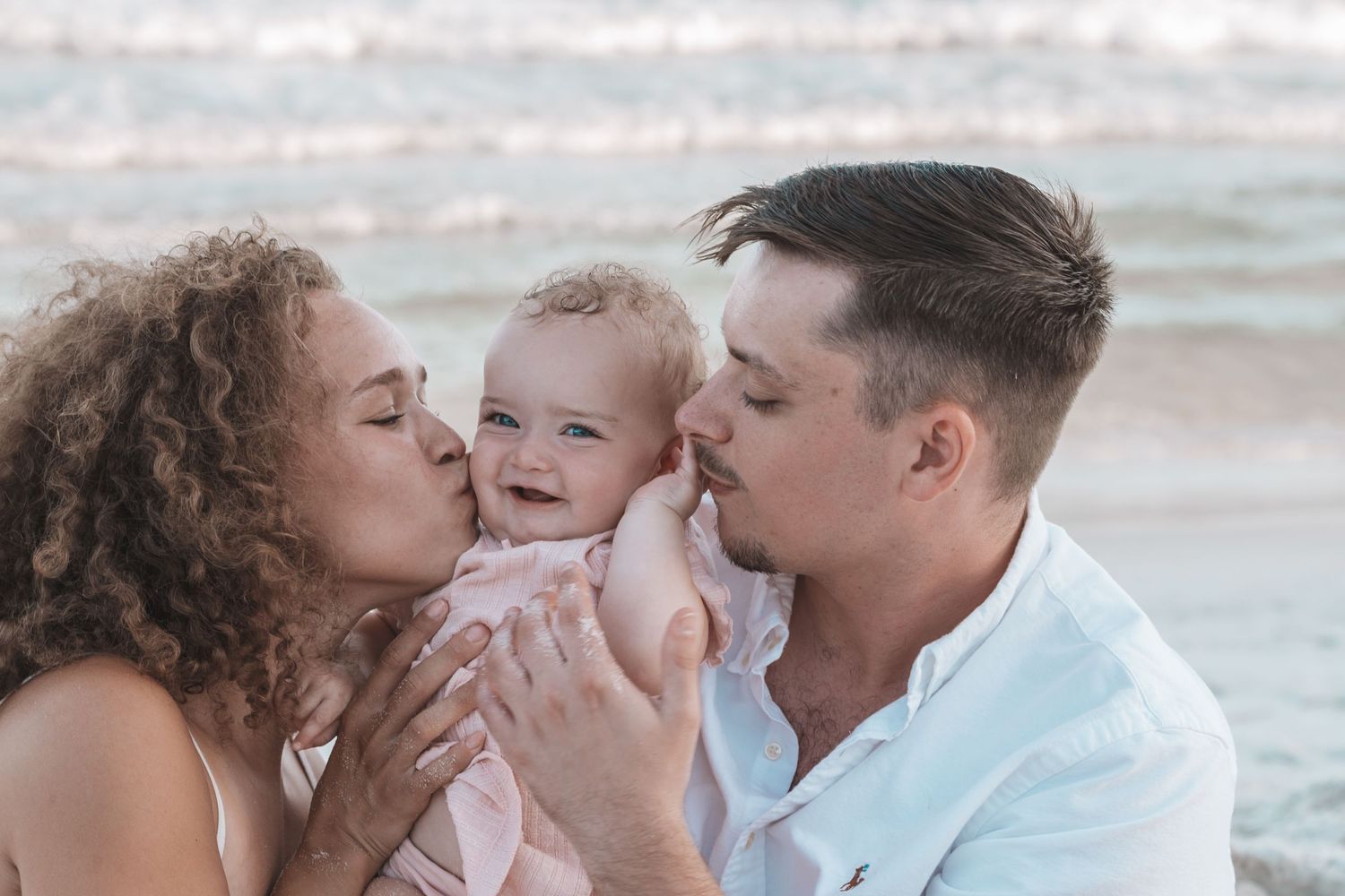 Parents kissing their smiling baby on the cheeks at a beach, family love moment.