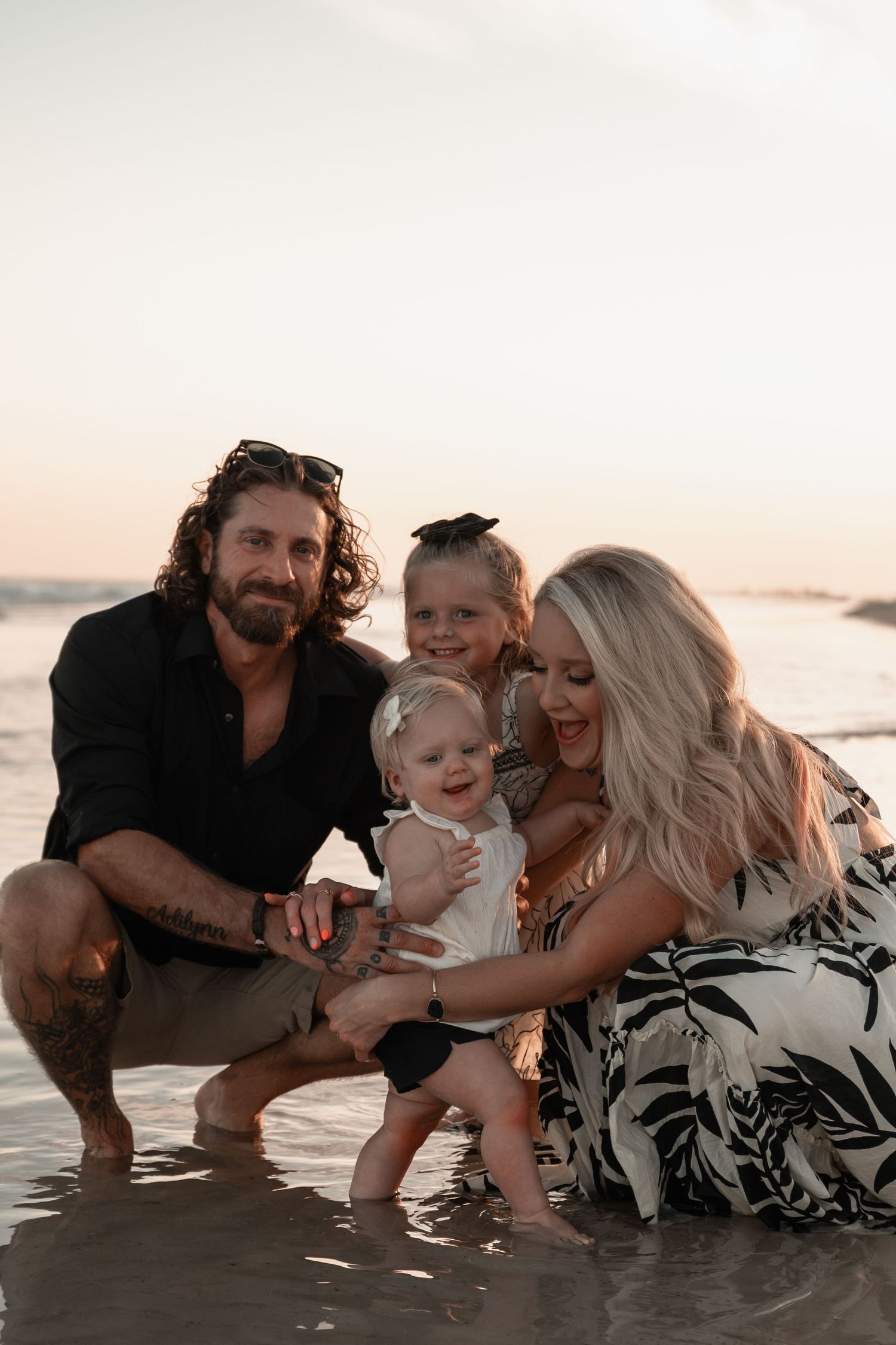 Happy family of four posing together at the water's edge on a beach at sunset.