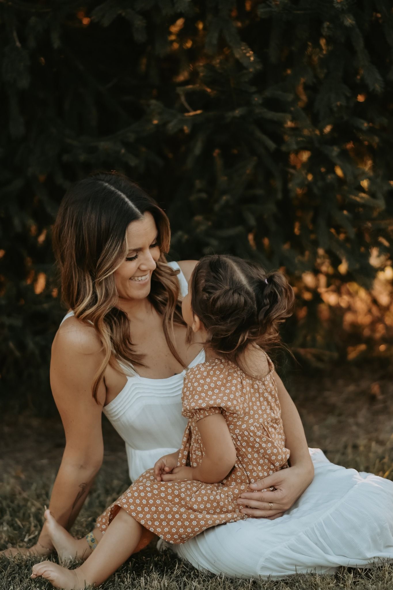 Loving mother in a white dress smiling warmly at her young daughter outdoors at golden hour.