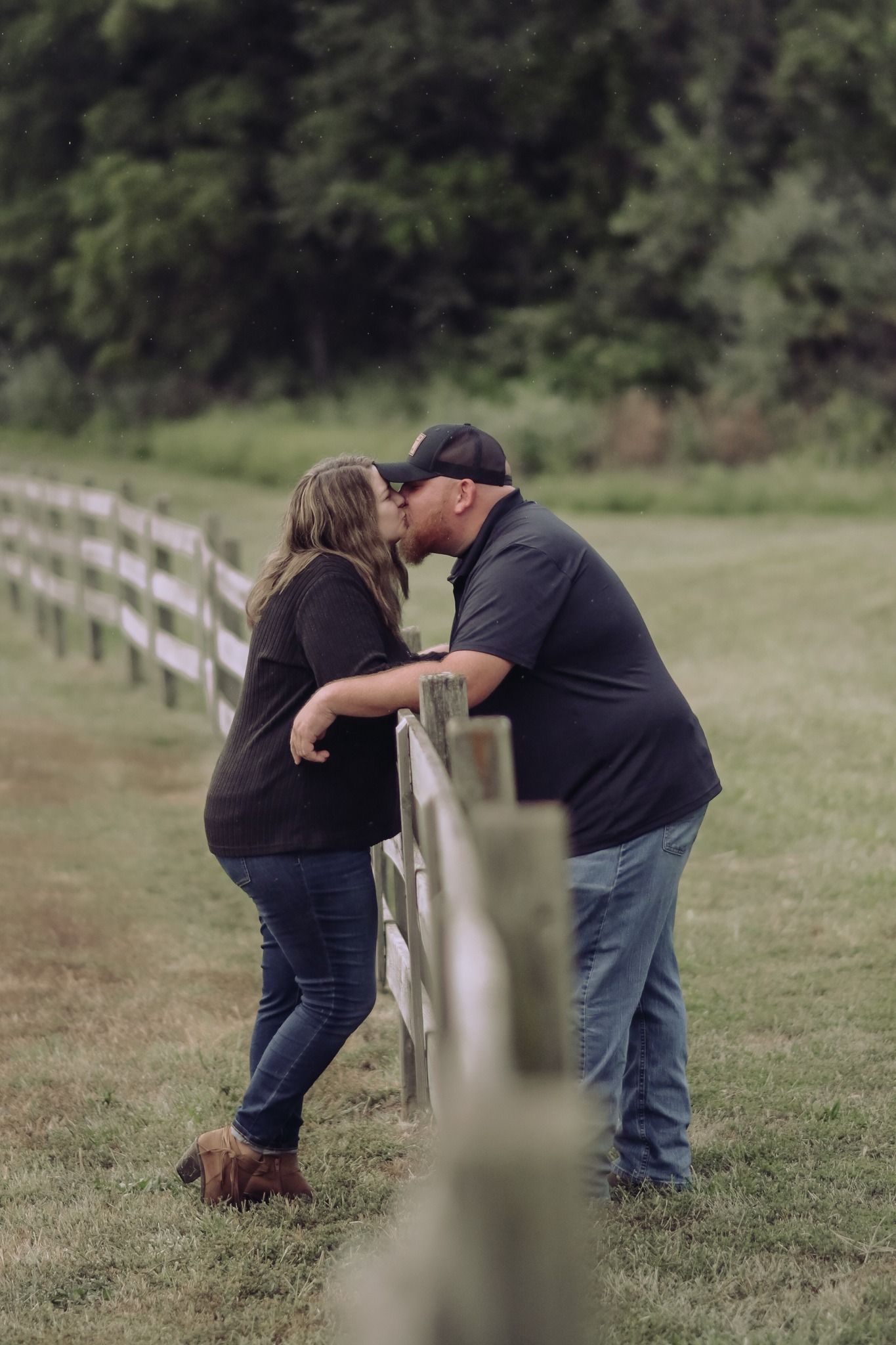 Couple sharing a romantic kiss leaning against a white wooden fence in a lush green countryside setting.