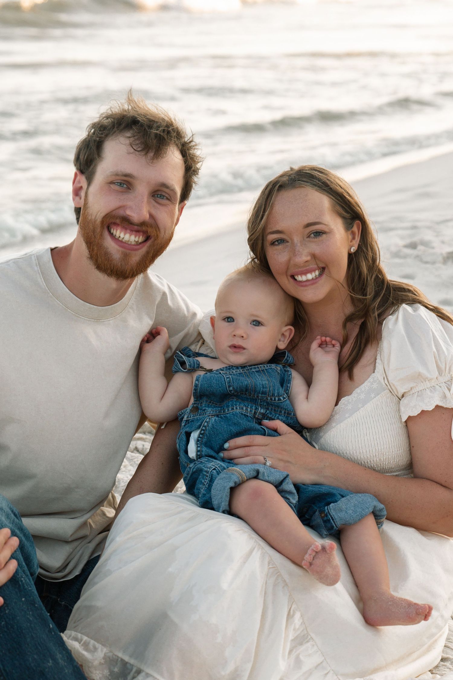 Happy young couple sitting on beach with baby in denim overalls, smiling at camera with ocean waves behind them.