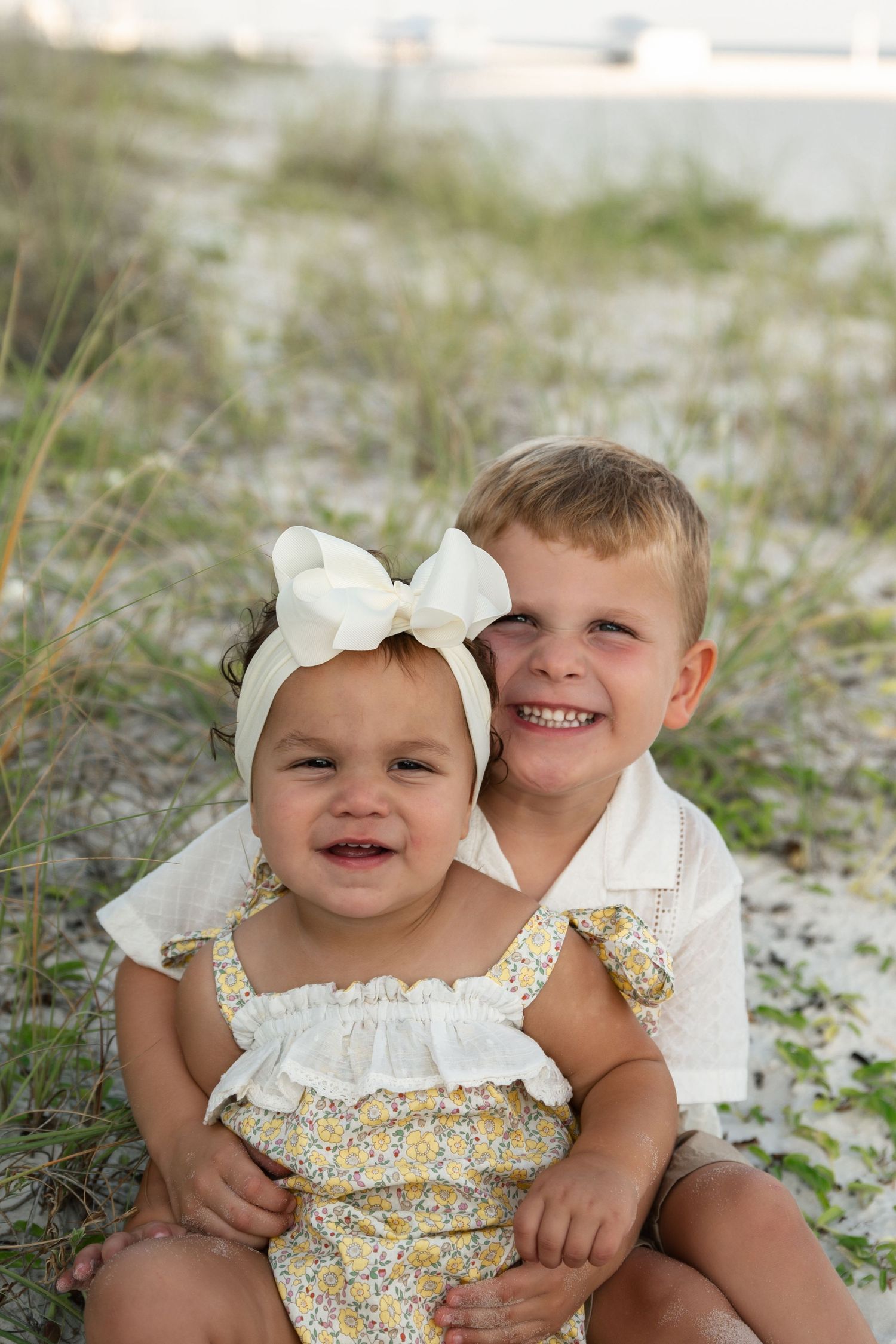 Two young siblings smile together on a sandy beach, girl in floral romper with bow headband, boy in white shirt.