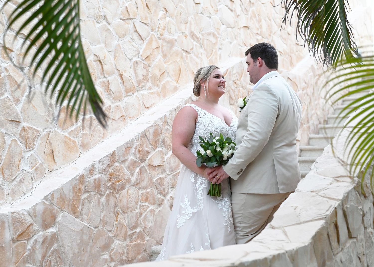 Newlywed couple sharing a romantic moment on stone steps surrounded by tropical palm leaves at a destination wedding.