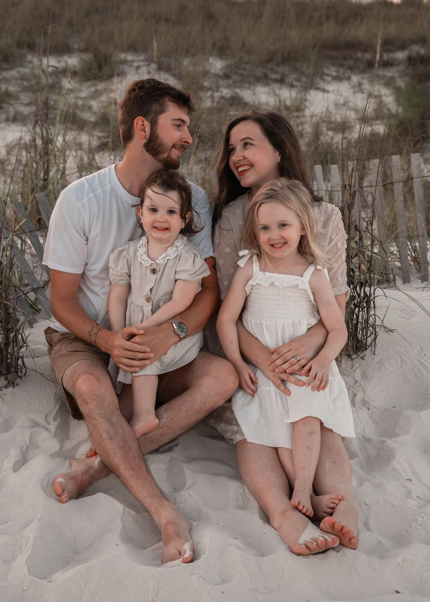 Happy family of four sitting on sandy beach dunes, parents with two young daughters in white outfits.