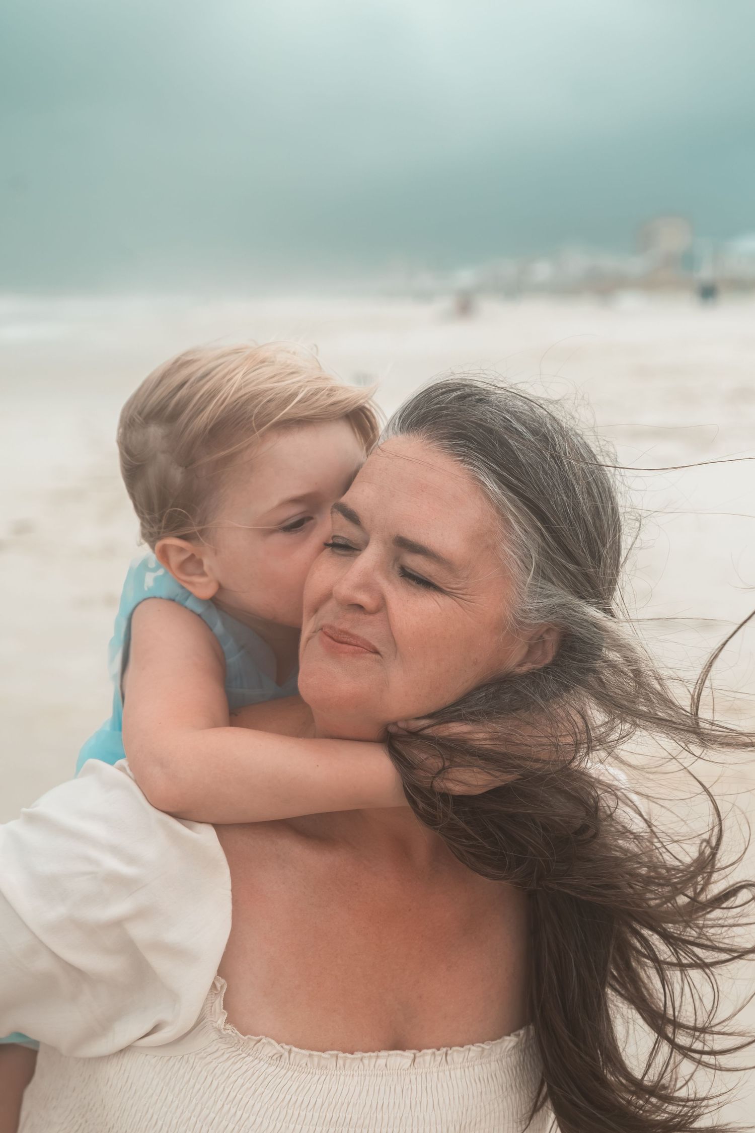 A grandmother lovingly holds her young grandchild on a breezy beach, sharing a tender cheek kiss moment.