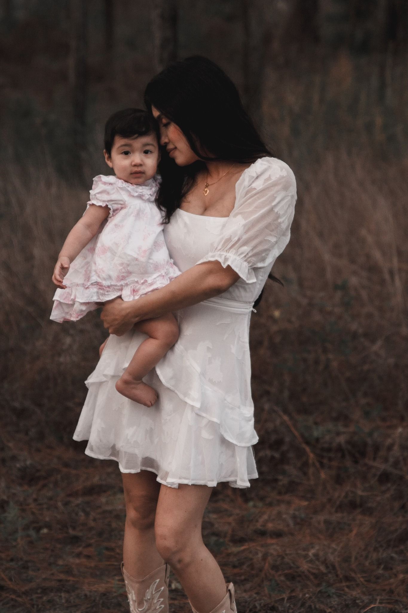 Asian woman in white dress lovingly holds baby girl in pink dress in wooded setting.