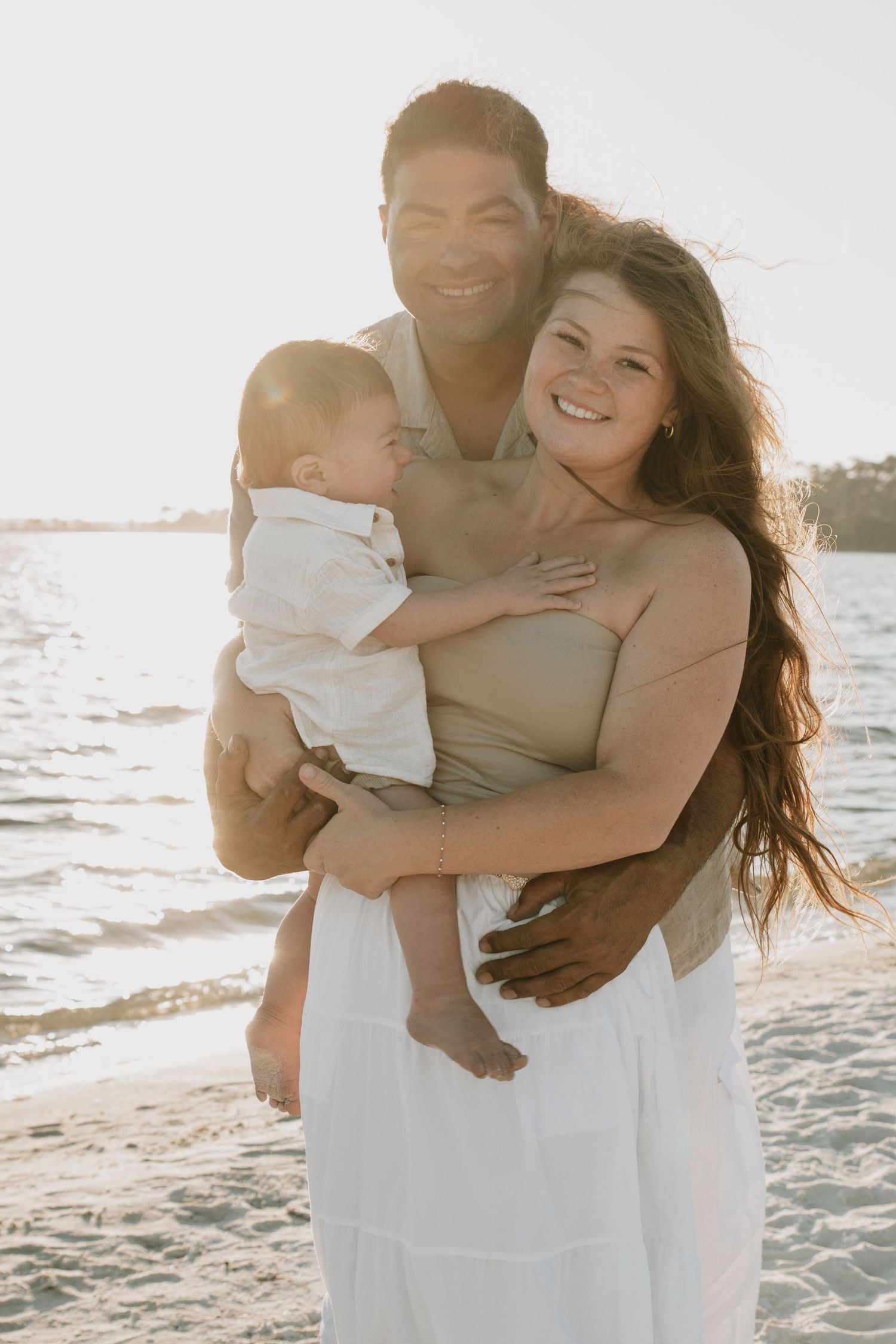 Smiling family of three poses together on a beach at sunset, parents holding their baby boy.
