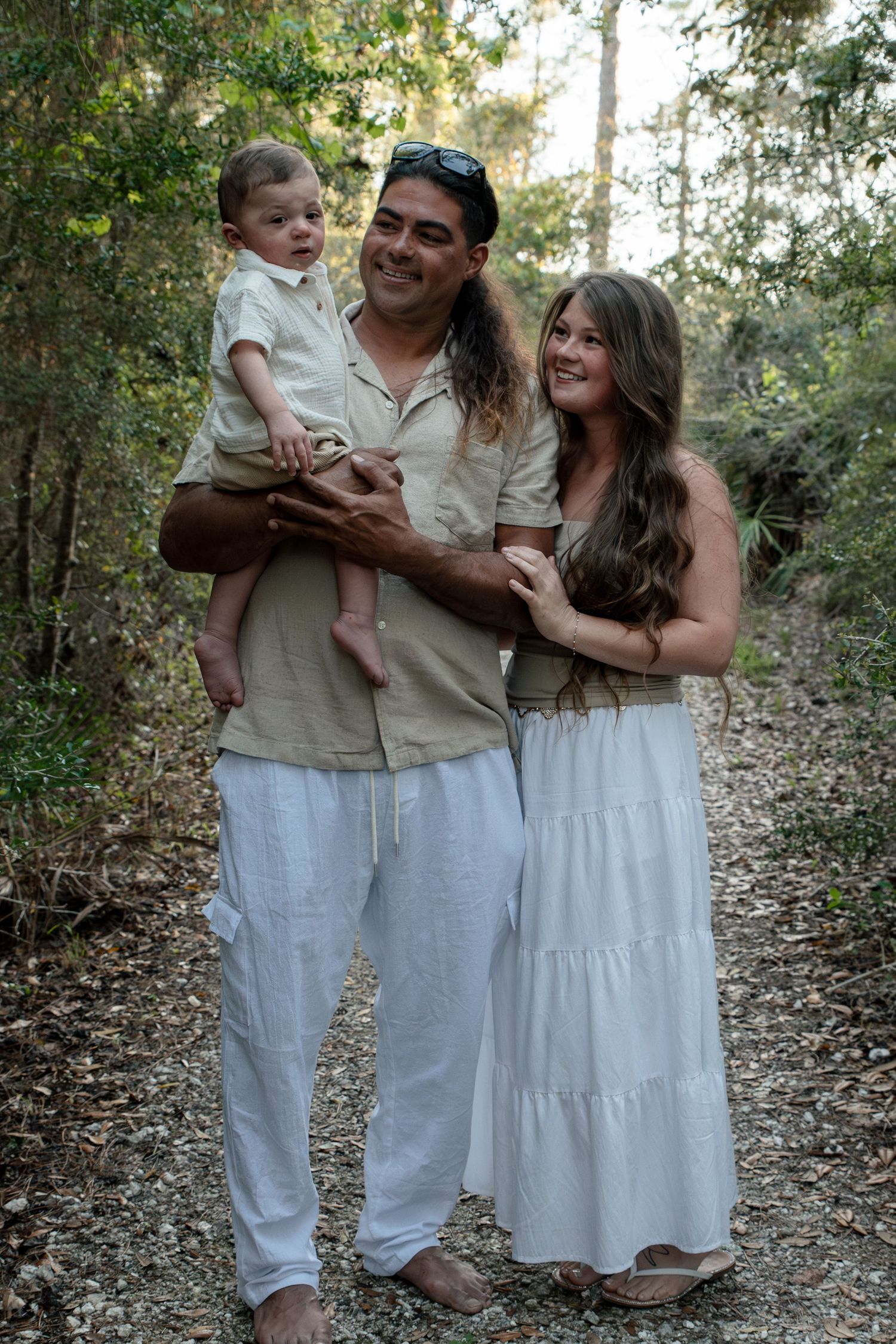 Smiling Hispanic family of three, parents holding baby, posing on a sunlit wooded path.