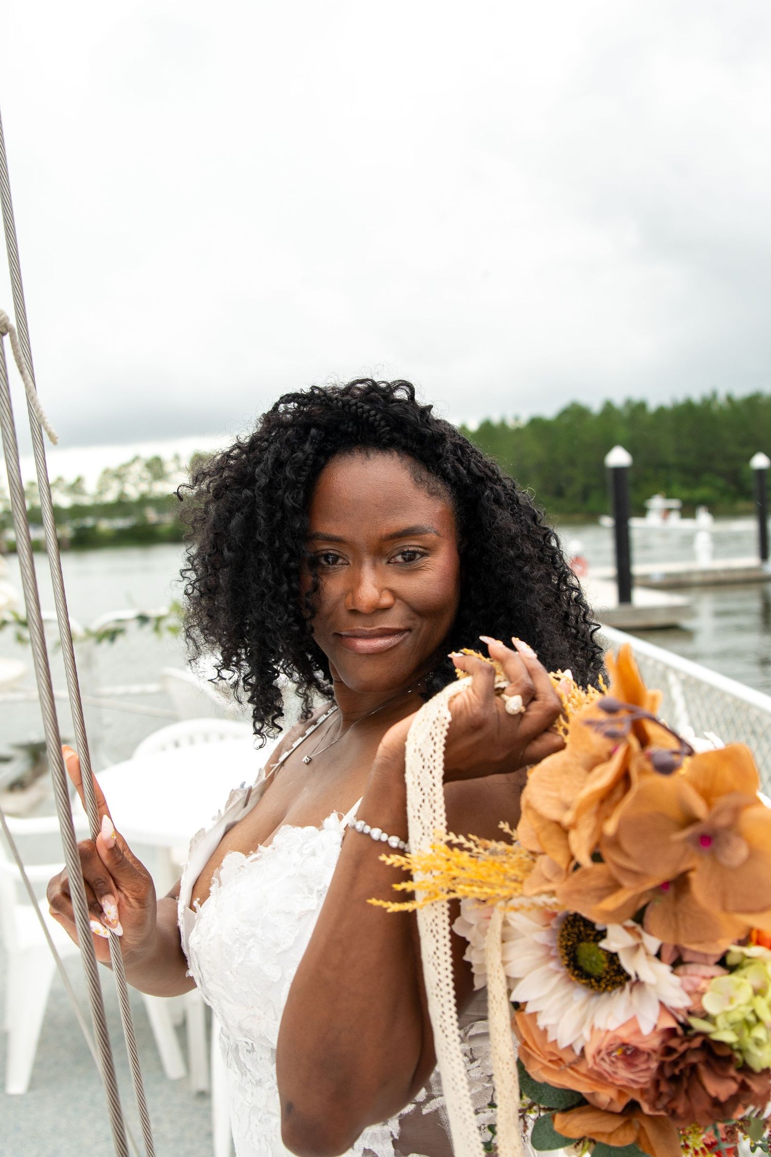 A Black bride in a white lace dress holds an autumn bouquet of orange and pink flowers on a waterfront deck.