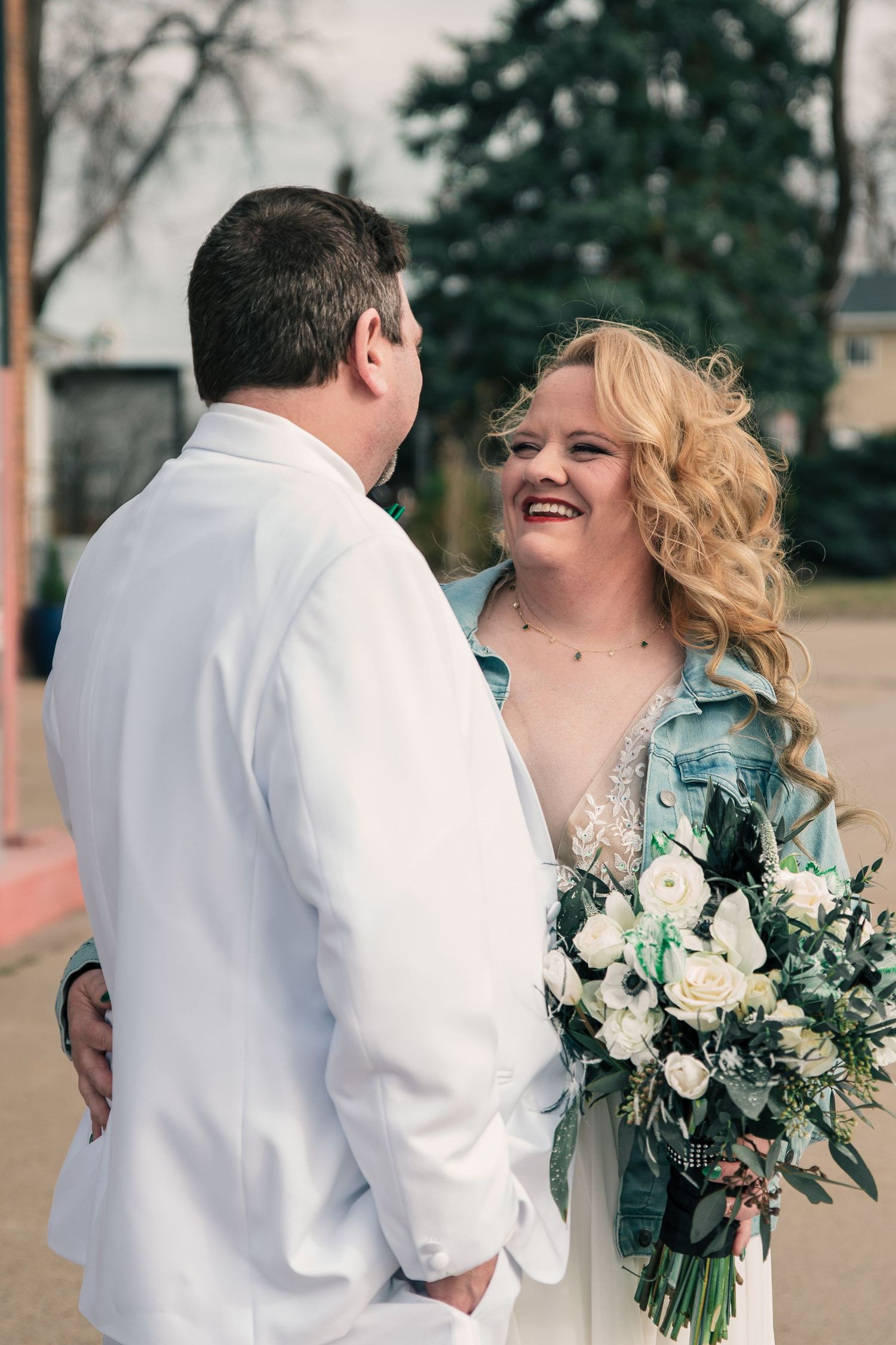 A laughing blonde bride in a denim jacket holds a white rose bouquet while gazing at her groom.