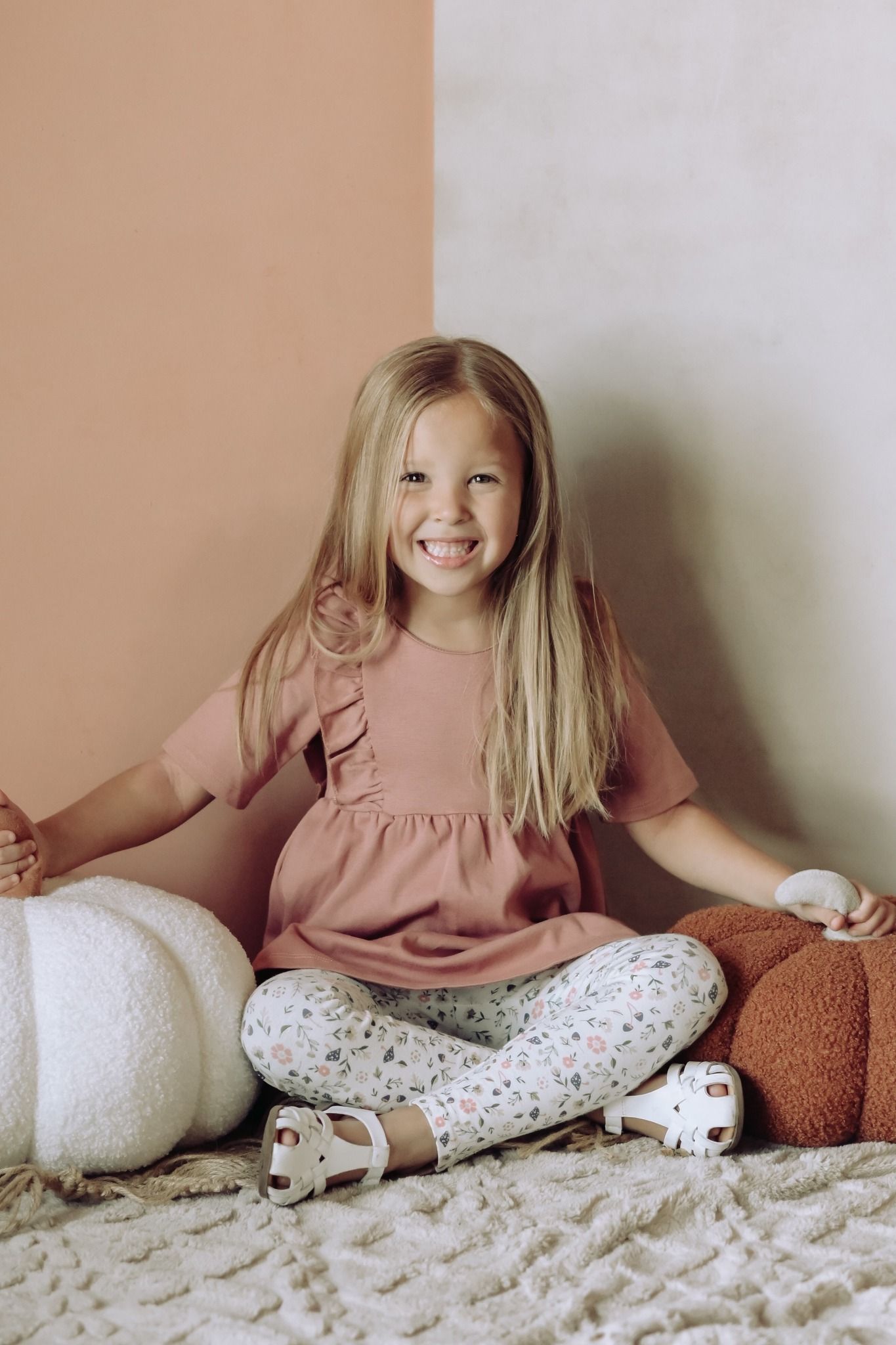 A cheerful blonde girl in a pink top and floral leggings sits cross-legged smiling at camera.