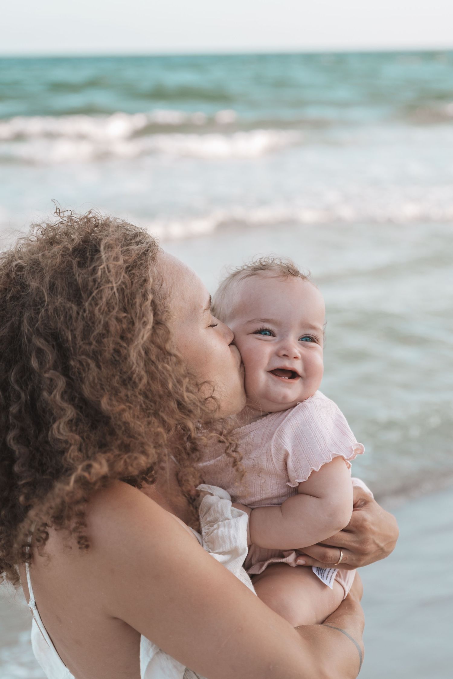 A curly-haired mother kissing her smiling baby girl on the cheek at the beach.