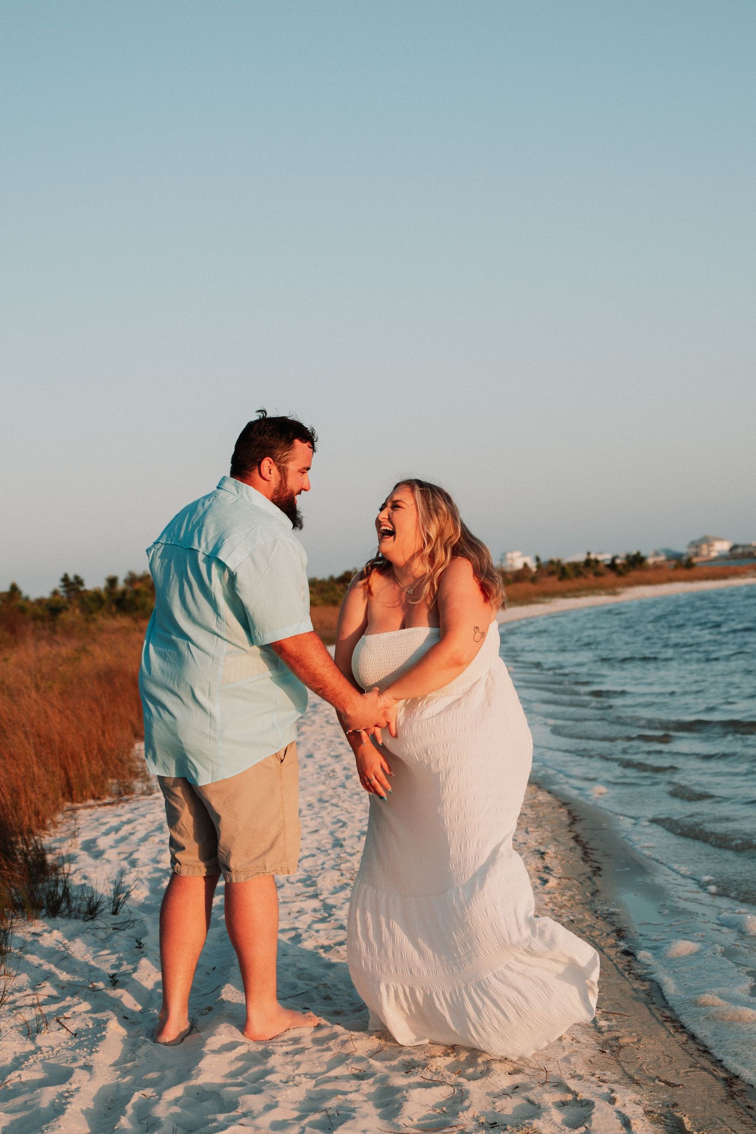 Laughing couple holds hands and dances together on a sandy beach at golden hour sunset.