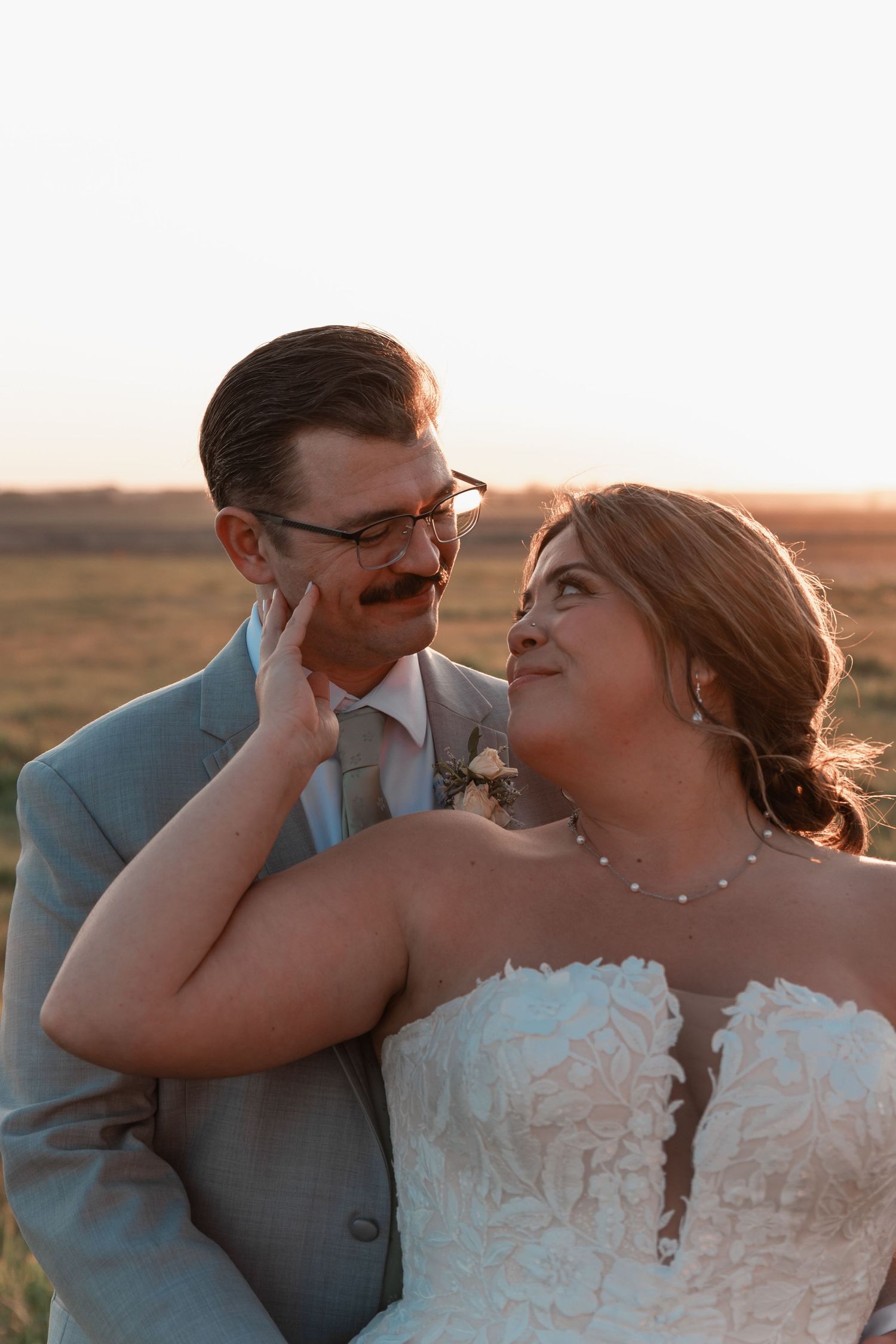 Bride tenderly touches grooms face as they gaze at each other in golden sunset light outdoors.