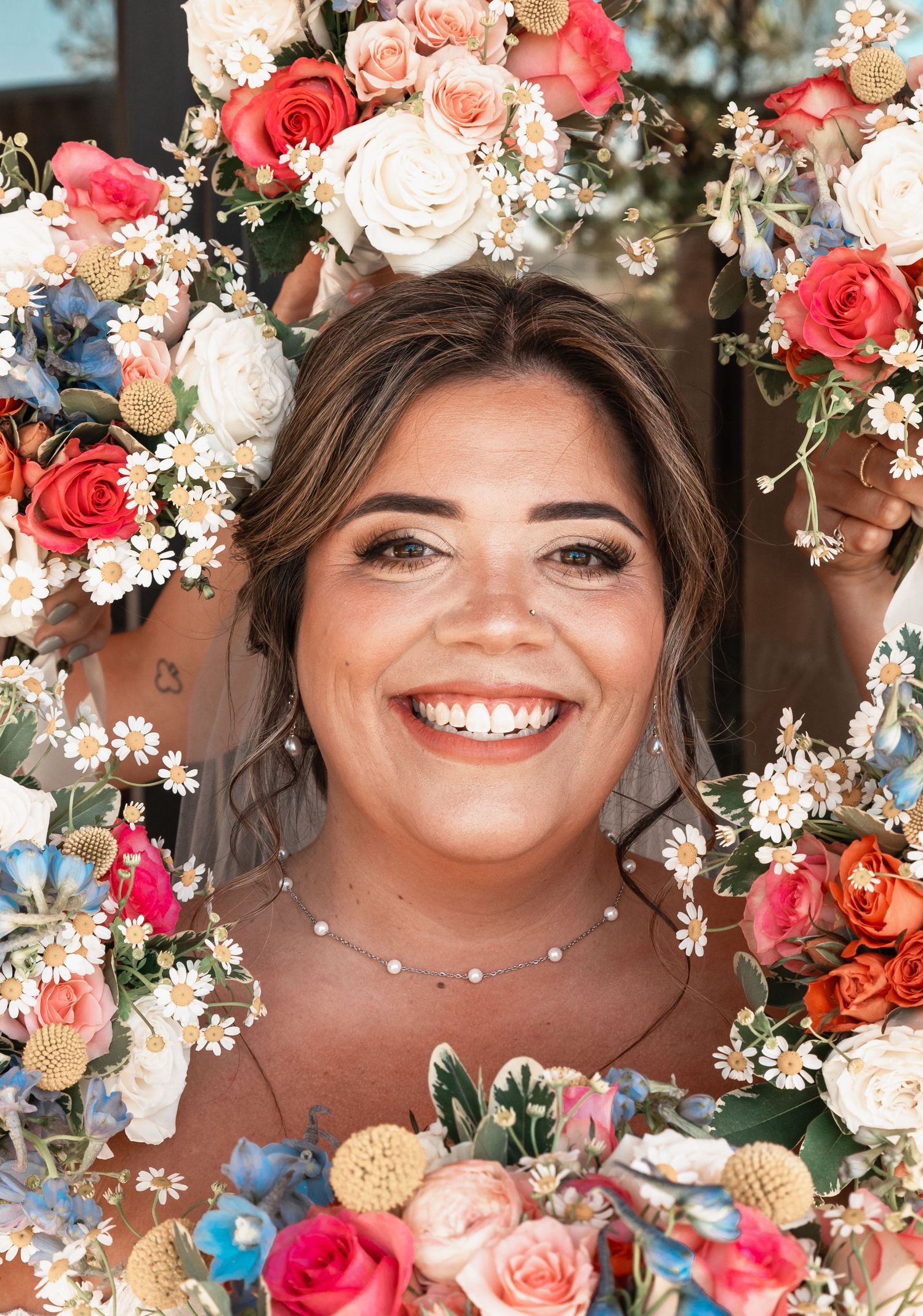 Smiling bride framed by colorful wedding bouquet featuring roses, daisies, and wildflowers.
