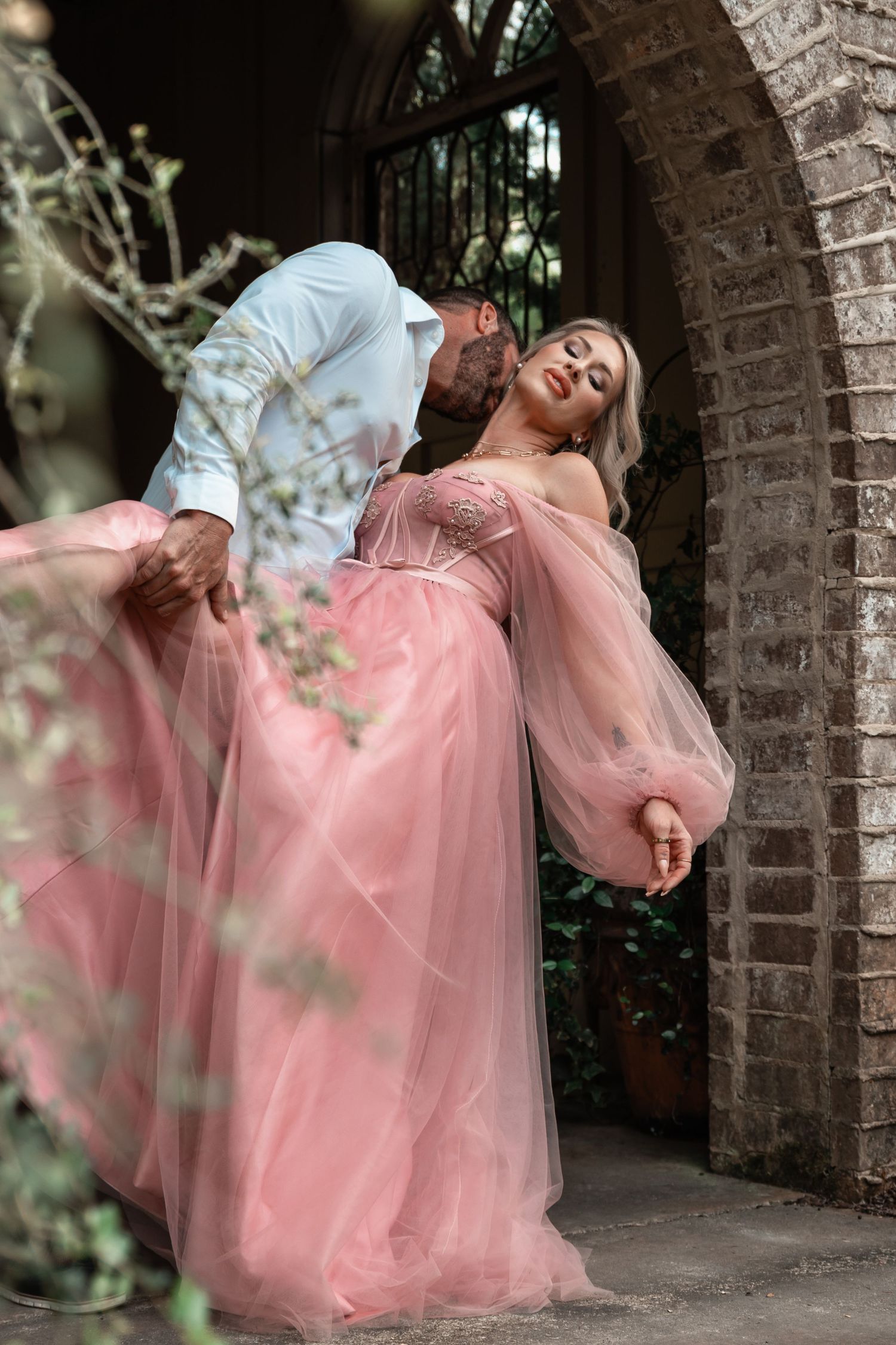 Couple shares an intimate moment under a brick arch, woman in pink off-shoulder tulle gown leans back gracefully.