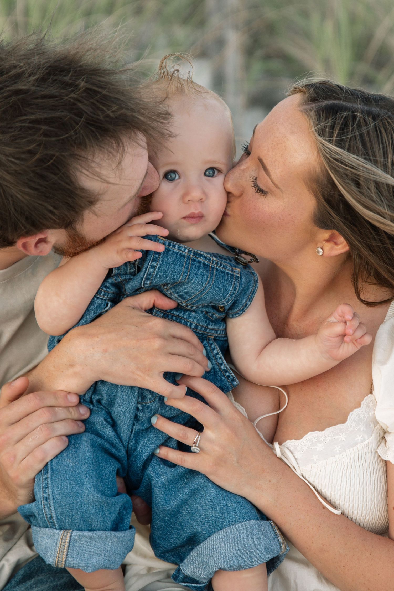 Two parents kissing their blue-eyed baby dressed in denim overalls during an outdoor family portrait session.