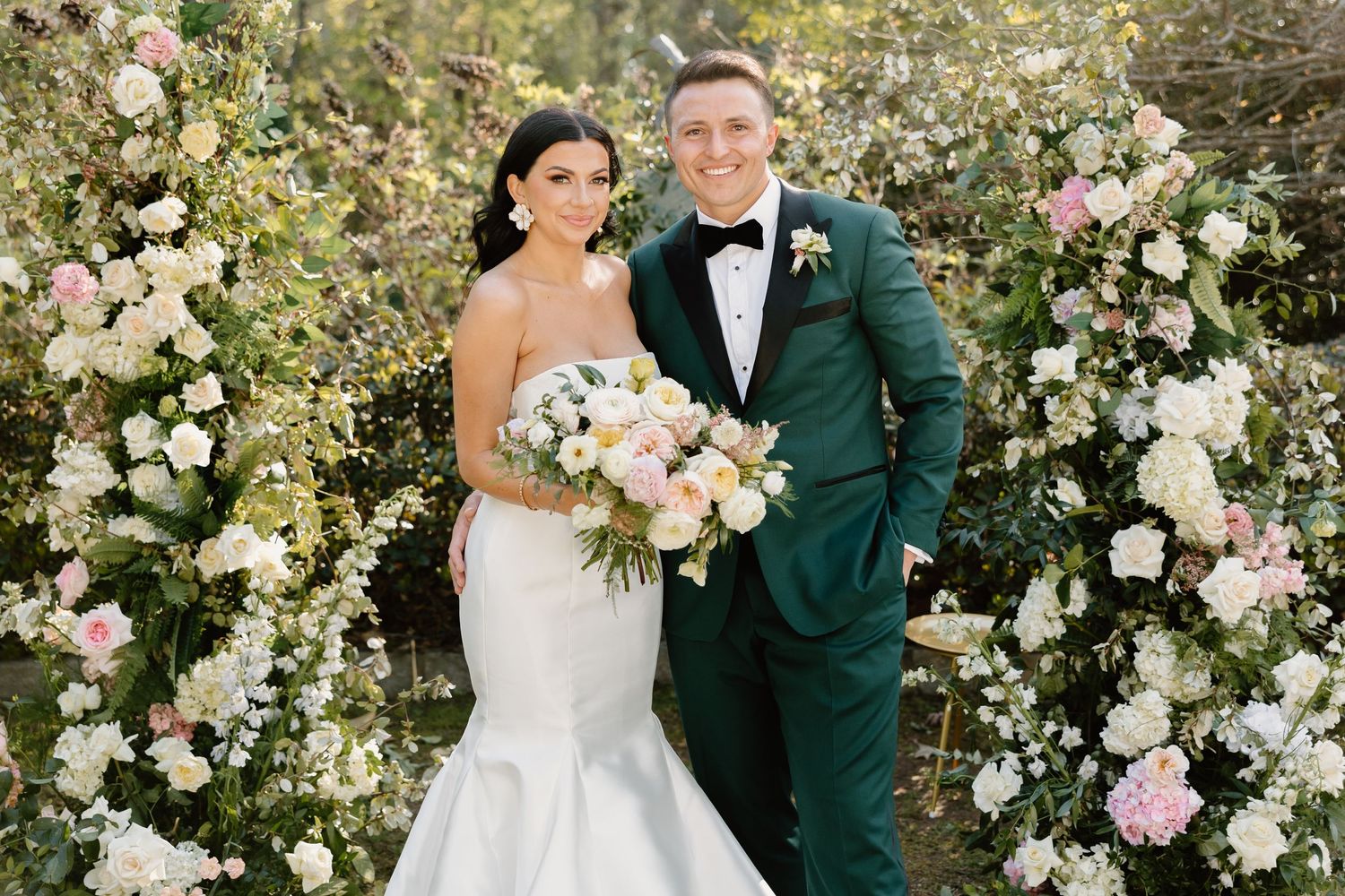 Bride with dark hair and trumpet skirt wedding gown holds pastel floral bouquet next to her groom in dark green tuxedo a