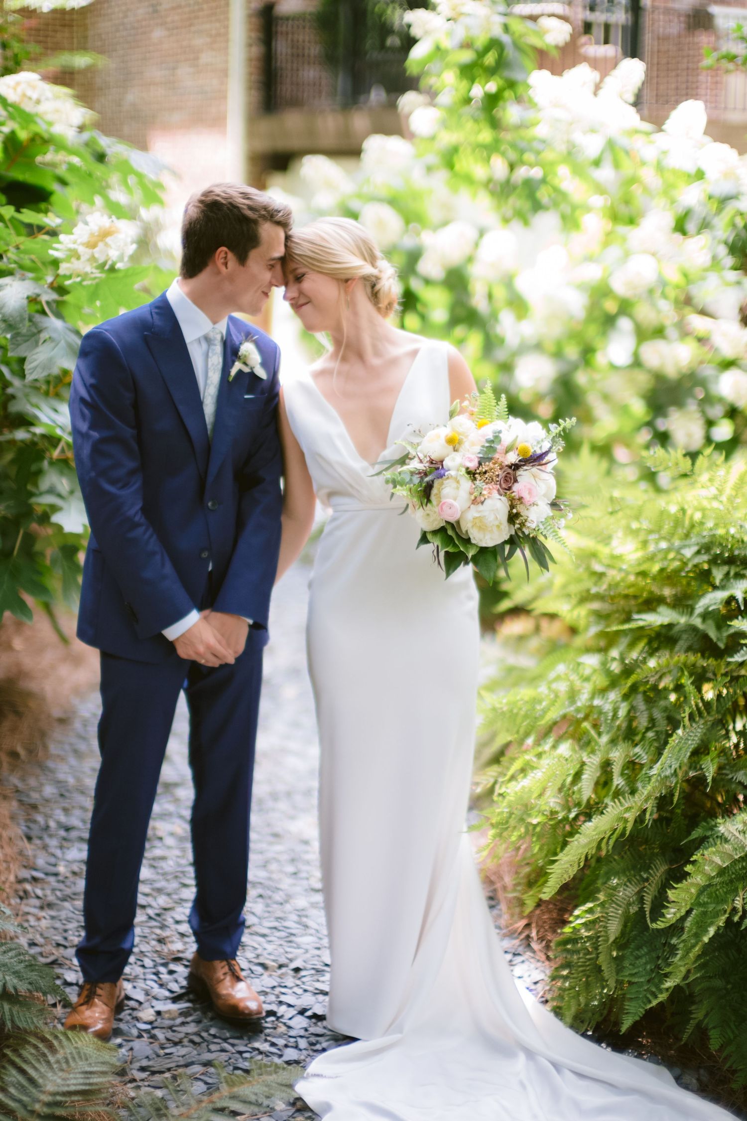 Bride with light hair in column-style wedding gown stands with groom in blue suit in at-home wedding coordinated by Gold