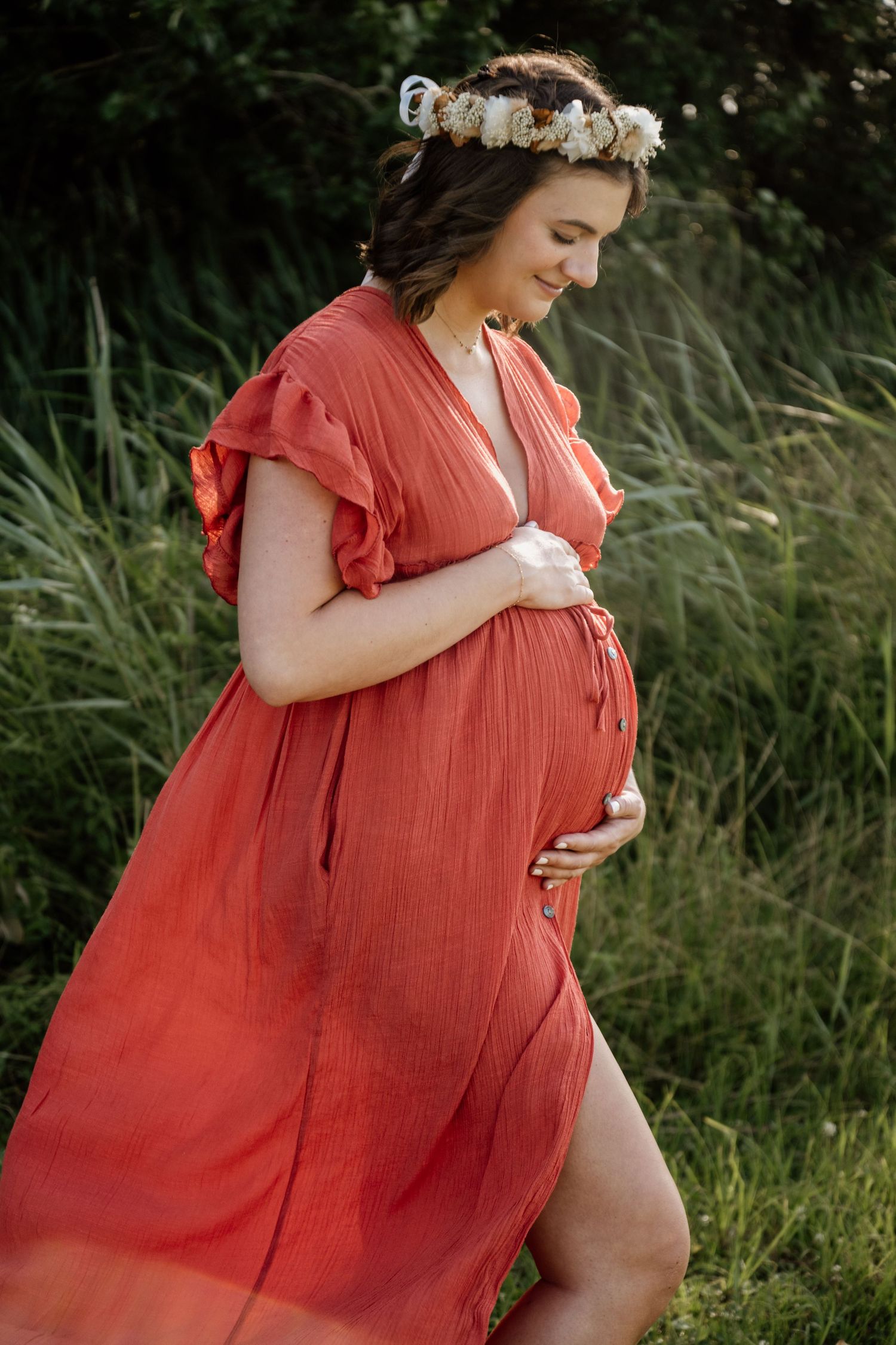 Pregnant woman in red dress and floral crown gently holds baby bump in nature.
