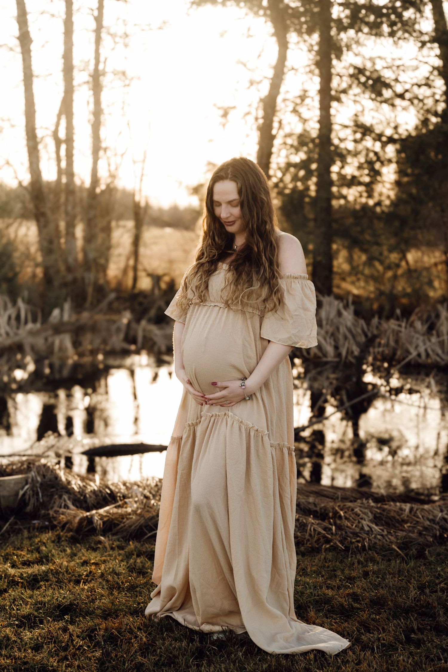 Pregnant woman in beige off-shoulder gown holds belly near golden sunset reflecting pond.