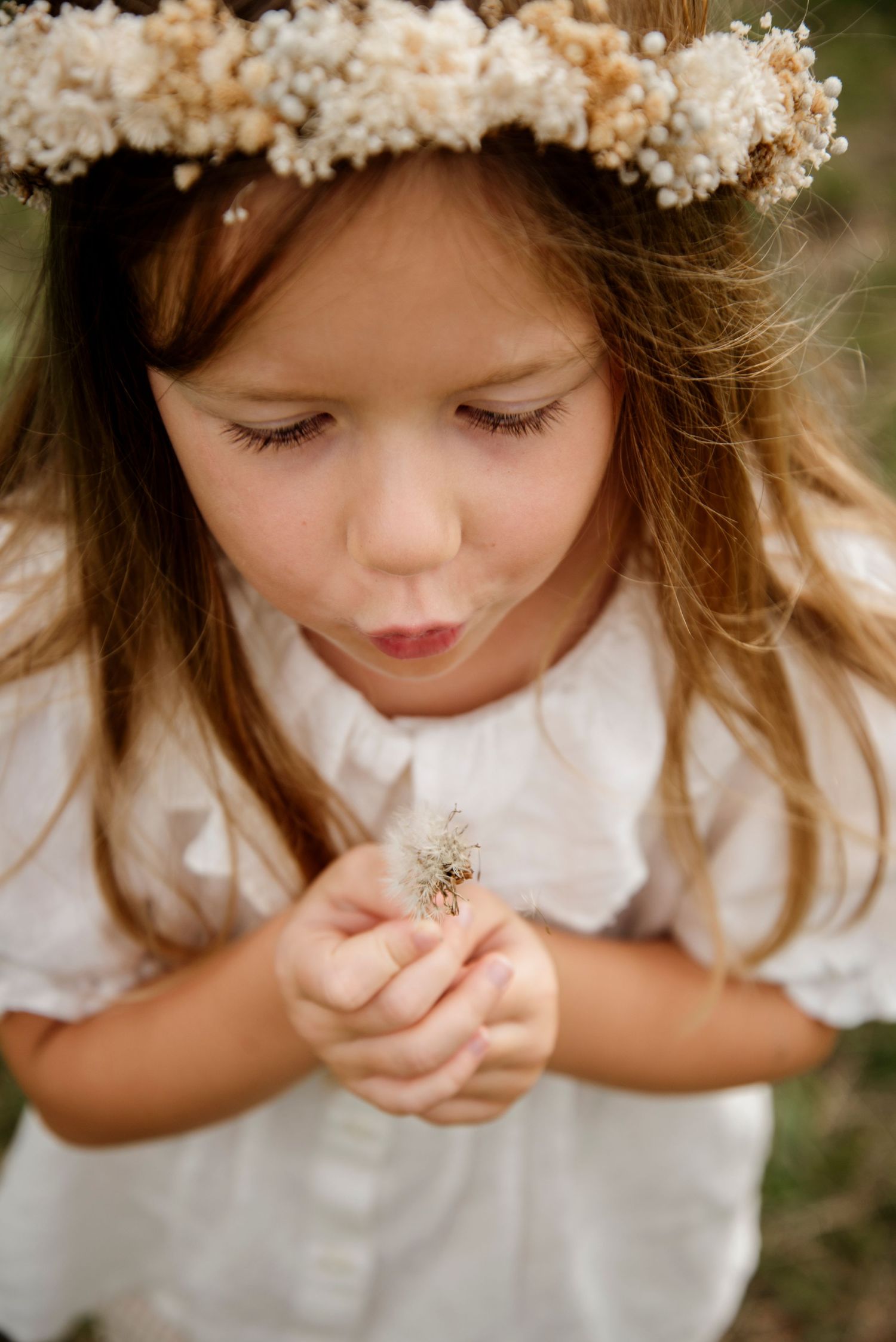 A young girl wearing a floral crown blows a dandelion while wearing a white dress outdoors.
